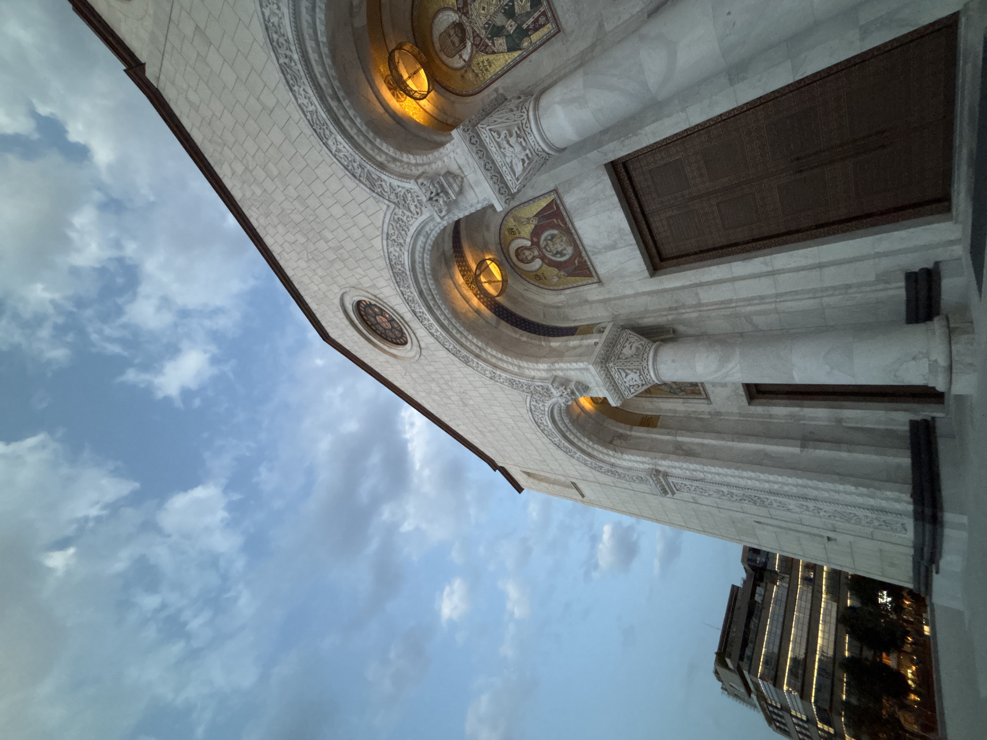 The image captures a low-angle view of the facade of the Cathedral of Saint Sava in Belgrade, Serbia, during twilight. The cathedral's white stone exterior, adorned with intricate carvings and arched entrances, is illuminated by warm artificial lights from within. Mosaic depictions of religious figures are visible above the arched doorways. Above the cathedral, a cloudy sky with patches of blue dominates the upper portion of the frame. To the left of the cathedral, a modern, multi-story building with a distinctive stepped facade and illuminated windows is visible, contrasting with the traditional architecture of the church. The overall scene suggests an urban environment in the evening, highlighting the juxtaposition of historical and contemporary structures.