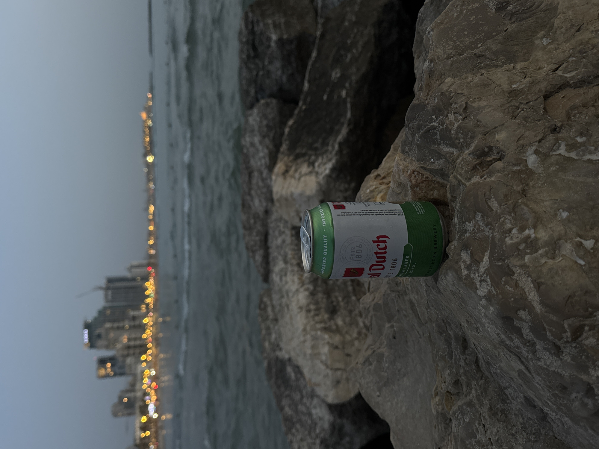 The image depicts a can of Royal Dutch beer resting on a rocky surface, possibly a breakwater. In the background is the coastline of Tel Aviv, Israel at night, with city lights blurred into streaks of yellow and white. The sea is visible between the rocks and the cityscape, under a gray sky.