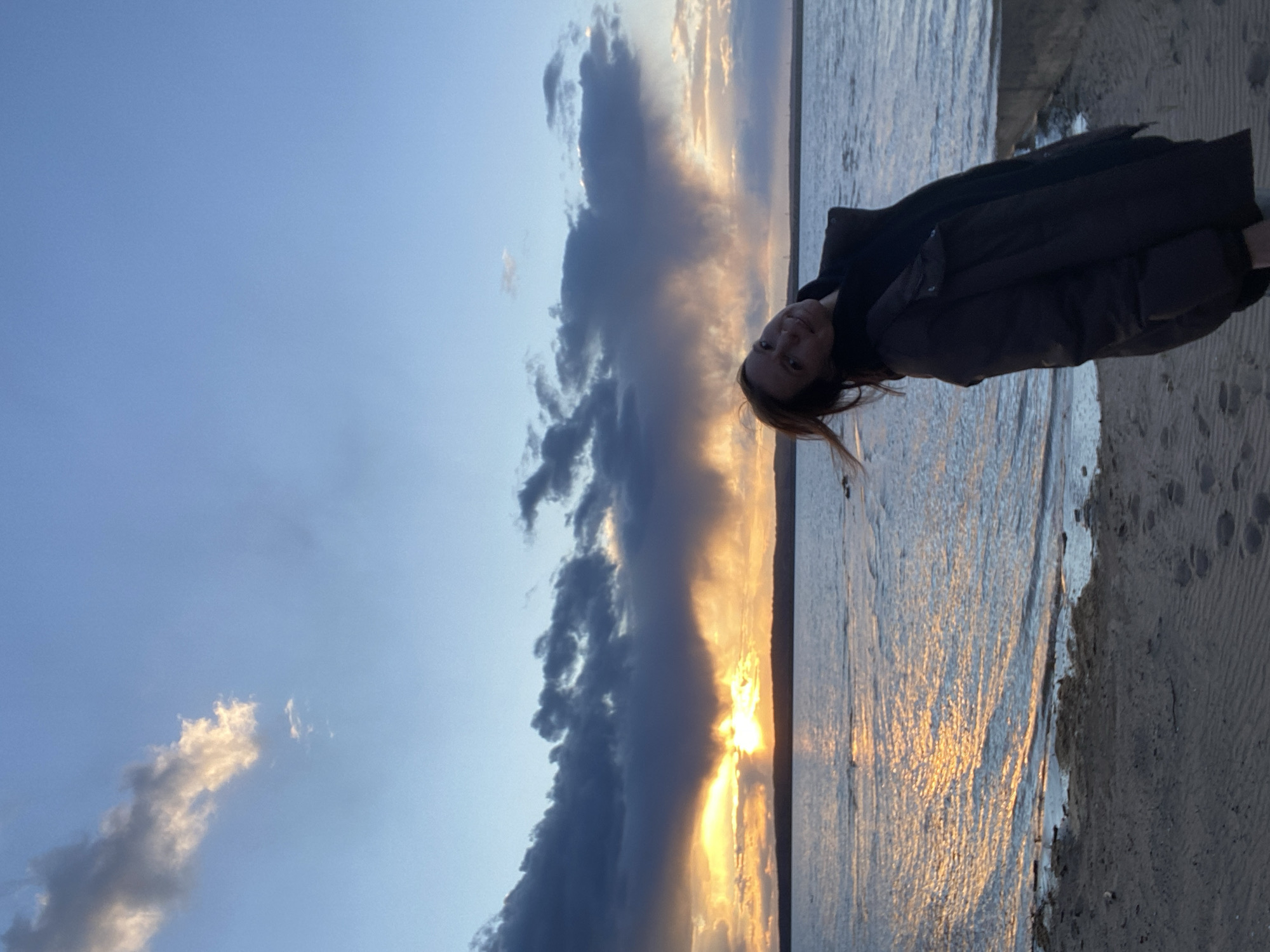 The image depicts a woman standing at the edge of a body of water, likely a river or a large lake, during sunset. The location is identified as Chaikovsky, Russia. The sky is a mix of blue and dramatic, dark clouds tinged with golden light from the setting sun. The sun's light reflects on the water's surface. The woman is wearing a dark coat. The ground she stands on appears to be sandy or gravelly, indicating a beach or riverbank.