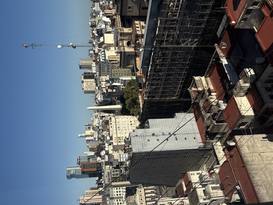 A cityscape under a clear blue sky features densely packed buildings. A tall antenna stands out against the horizon. The Obelisco landmark is visible among the structures. Scaffolding covers the side of a building under construction. Red rooftops and varying architectural styles contribute to the urban scene. Utility wires crisscross in the foreground.