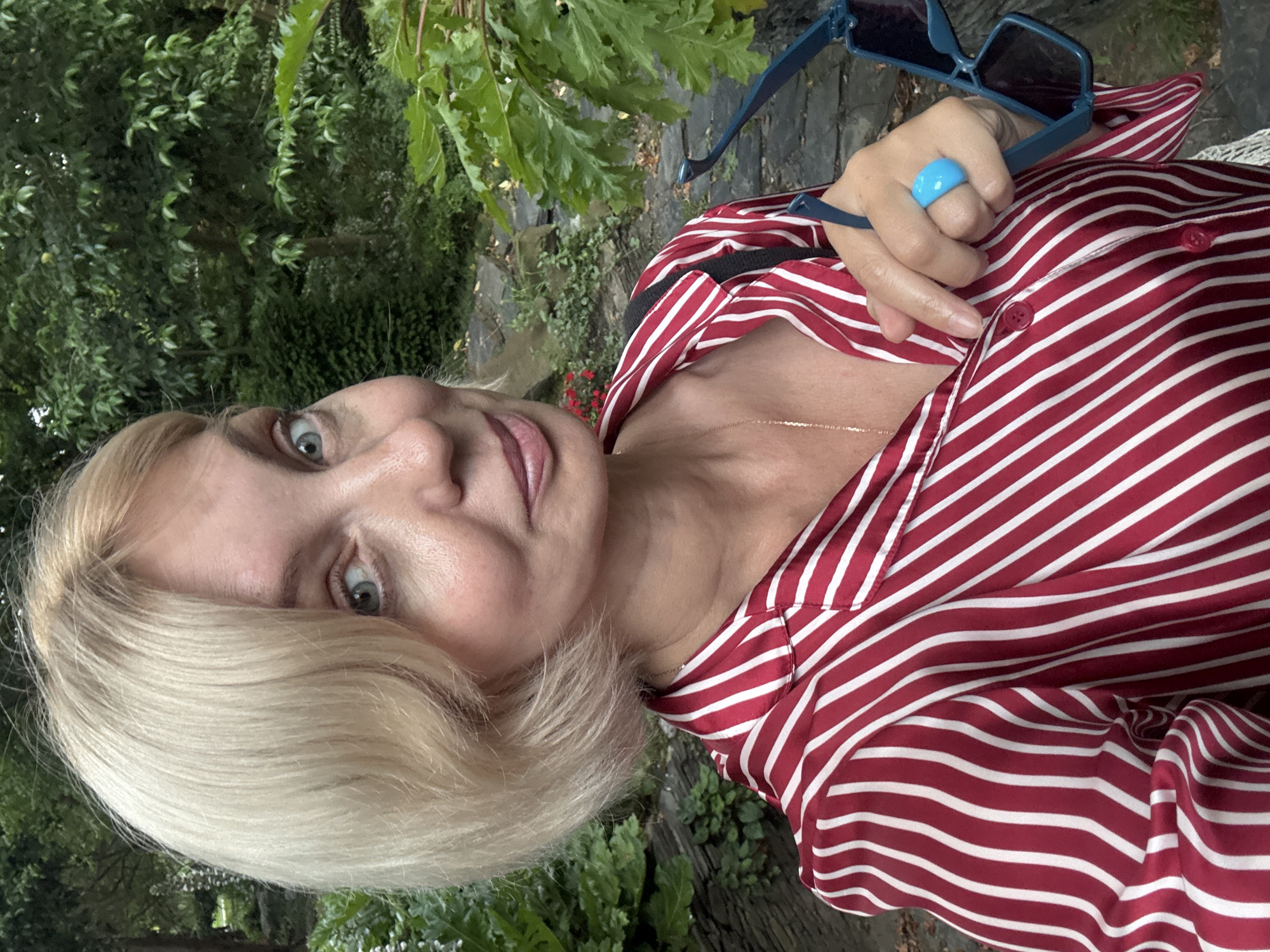 A blonde woman with short hair, wearing a red and white striped shirt, smiles as she holds sunglasses in her right hand. She is wearing a bright blue ring on her left ring finger. The background consists of lush green foliage, suggesting an outdoor, possibly park-like setting. The woman's location in the city of Sochi, Russia is unknown.