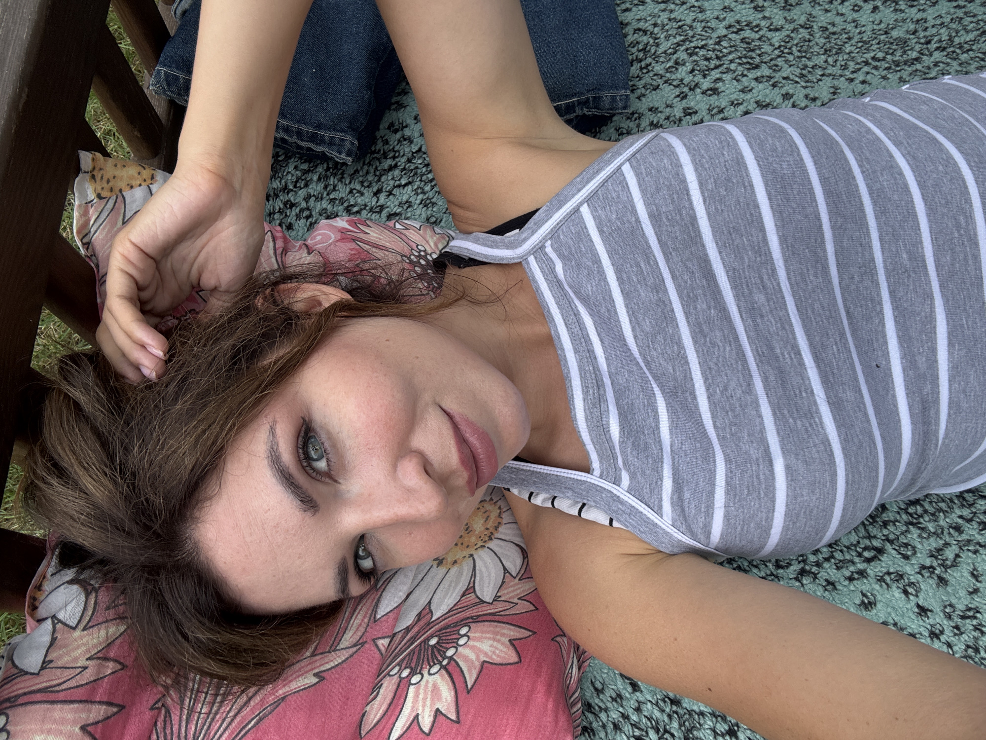 A woman with light brown hair and blue eyes is lying on her back on a patterned blanket outdoors. She is wearing a grey and white striped tank top and blue denim shorts. Her left arm is bent at the elbow, with her hand resting near her head. Her head is tilted back slightly, and her gaze is directed upwards. The background consists of grass and the edge of a wooden structure. The scene appears to be in a natural outdoor setting, possibly a park or a garden, within the city of Lozuvatka, Ukraine.