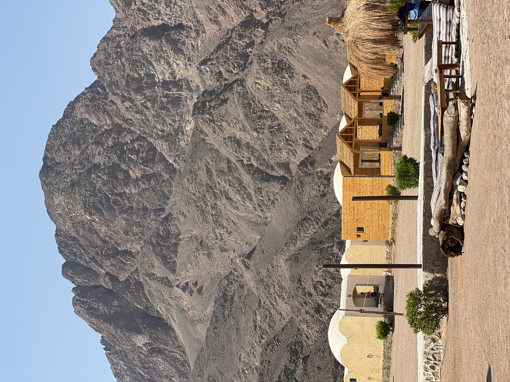 In Nueiba, Egypt, a settlement of small, wooden cabins with thatched roofs is nestled against the base of a vast, rocky mountain range. The cabins are arranged in a row, with a sandy courtyard in front of them. A few green bushes dot the landscape, and a thatched umbrella provides shade over a seating area with striped cushions. In the background, the imposing mountain dominates the sky, its rugged slopes a testament to the arid environment. A person is visible standing in the doorway of one of the buildings, partially obscured. The scene suggests a tranquil desert retreat.