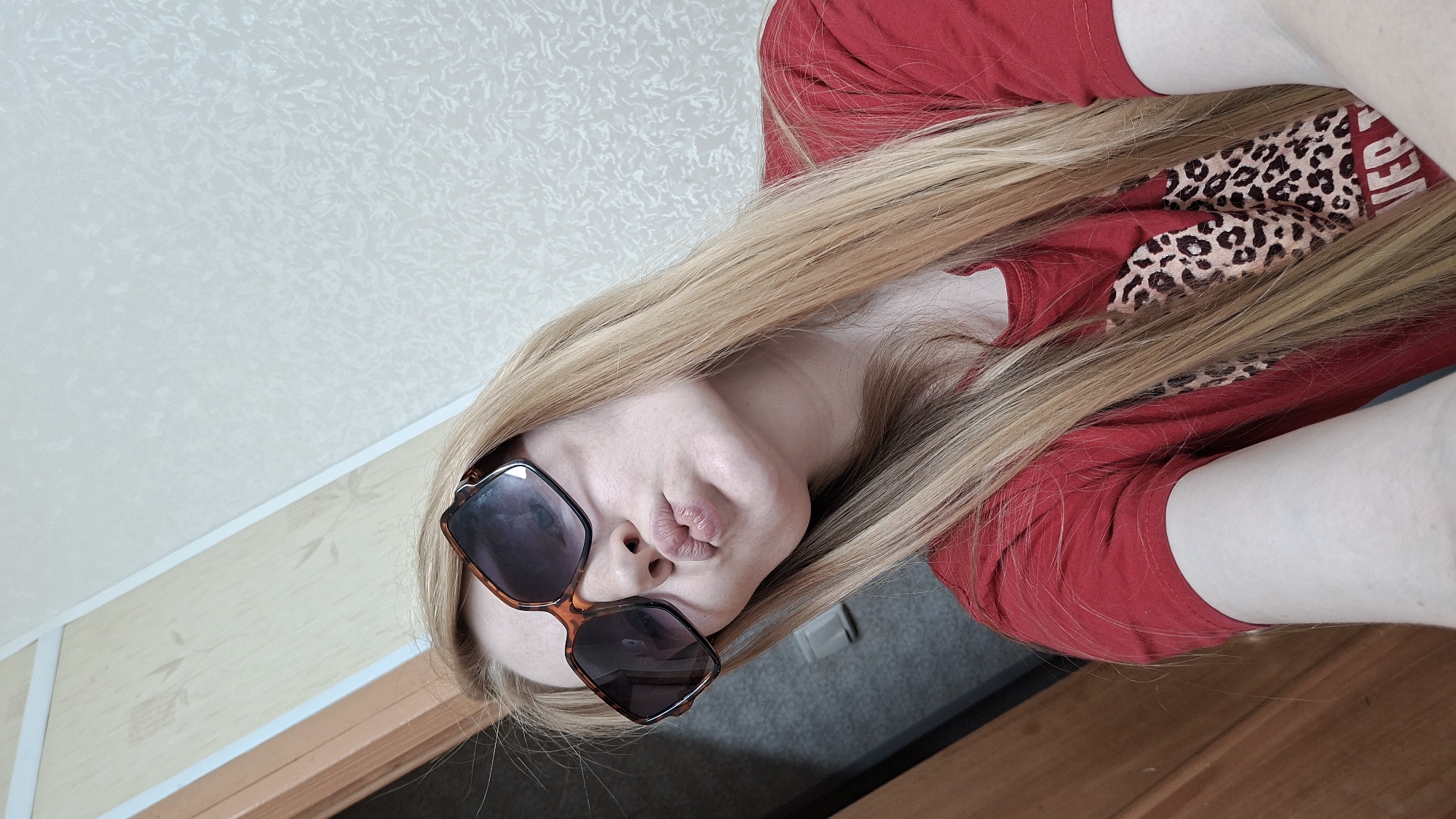 A young woman with long, blonde hair and sunglasses is taking a selfie. She is wearing a red t-shirt with a leopard print design. The background appears to be indoors, possibly in a home, with a doorframe and patterned wallpaper visible. The woman is puckering her lips, as if blowing a kiss. The image does not contain information about the city of Большой Букор, Russia.