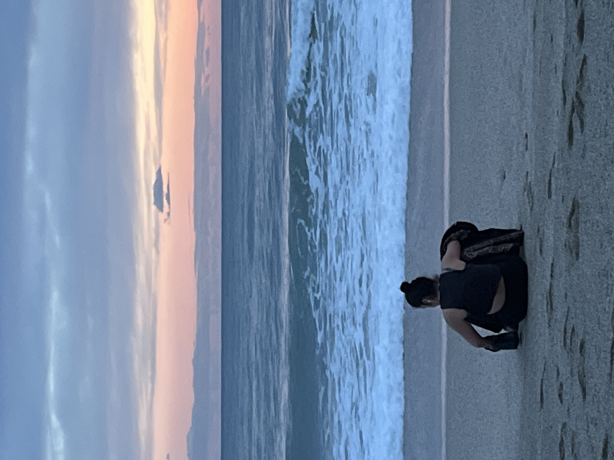 The image shows a woman crouching on a beach next to ocean waves. She is wearing a dark-colored shirt and pants. The sky displays shades of pink and blue. The scene is likely taken at dusk or dawn, with soft lighting. The location cannot be identified as Alanya, Turkey, based on the image alone.
