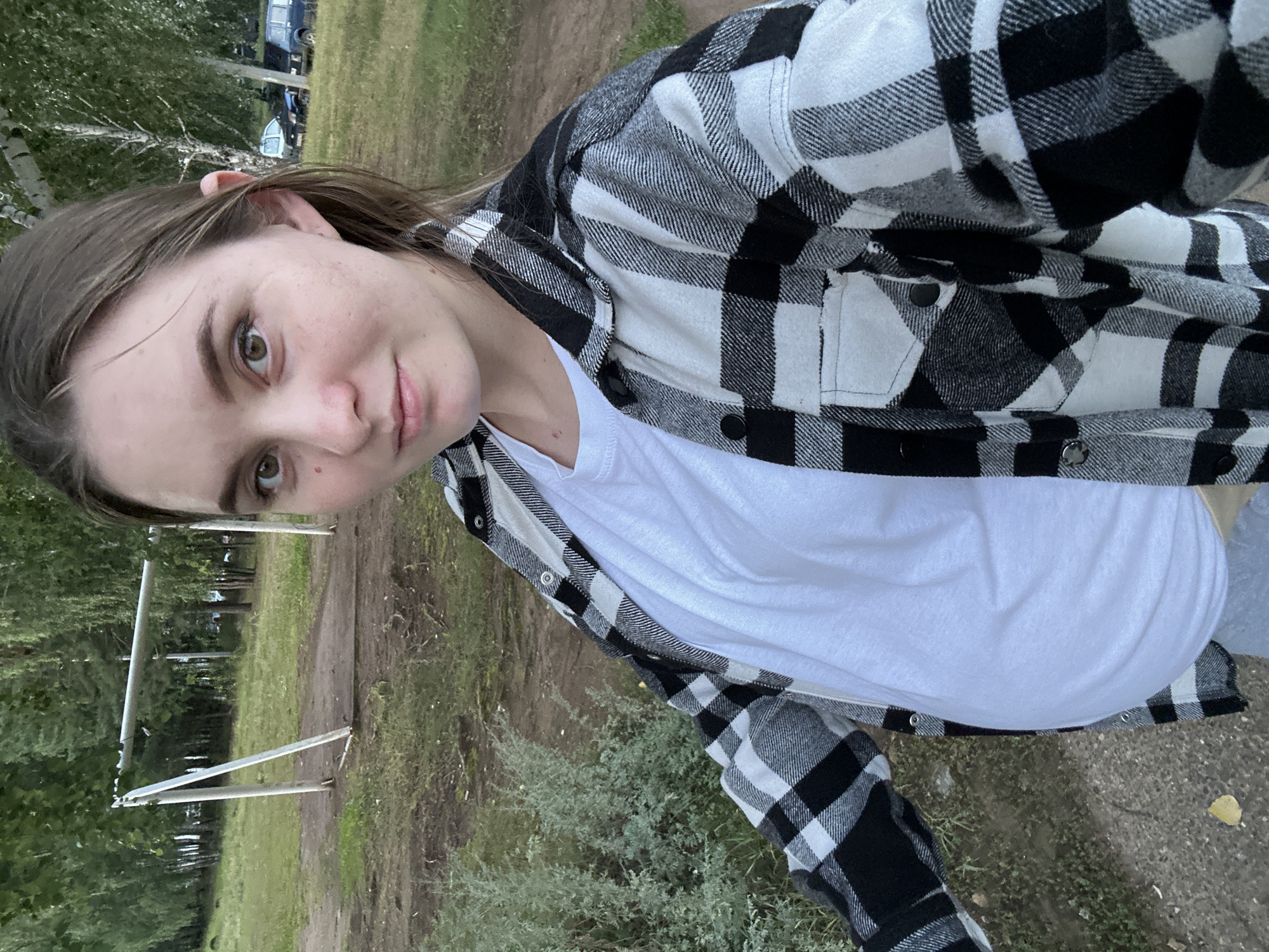 A young Caucasian woman with light brown hair pulled back and brown eyes is standing outdoors, taking a selfie. She is wearing a white t-shirt and a black and white plaid shirt unbuttoned over it. She has a light complexion with some freckles on her cheeks and a mole on her left cheek. She is smiling slightly. The background shows a grassy area with trees and a white soccer goal. A dirt path runs through the scene, and a few cars are visible in the distance amongst the trees. The image appears to have been taken in the city of Noviy, Russia, based on the provided information.