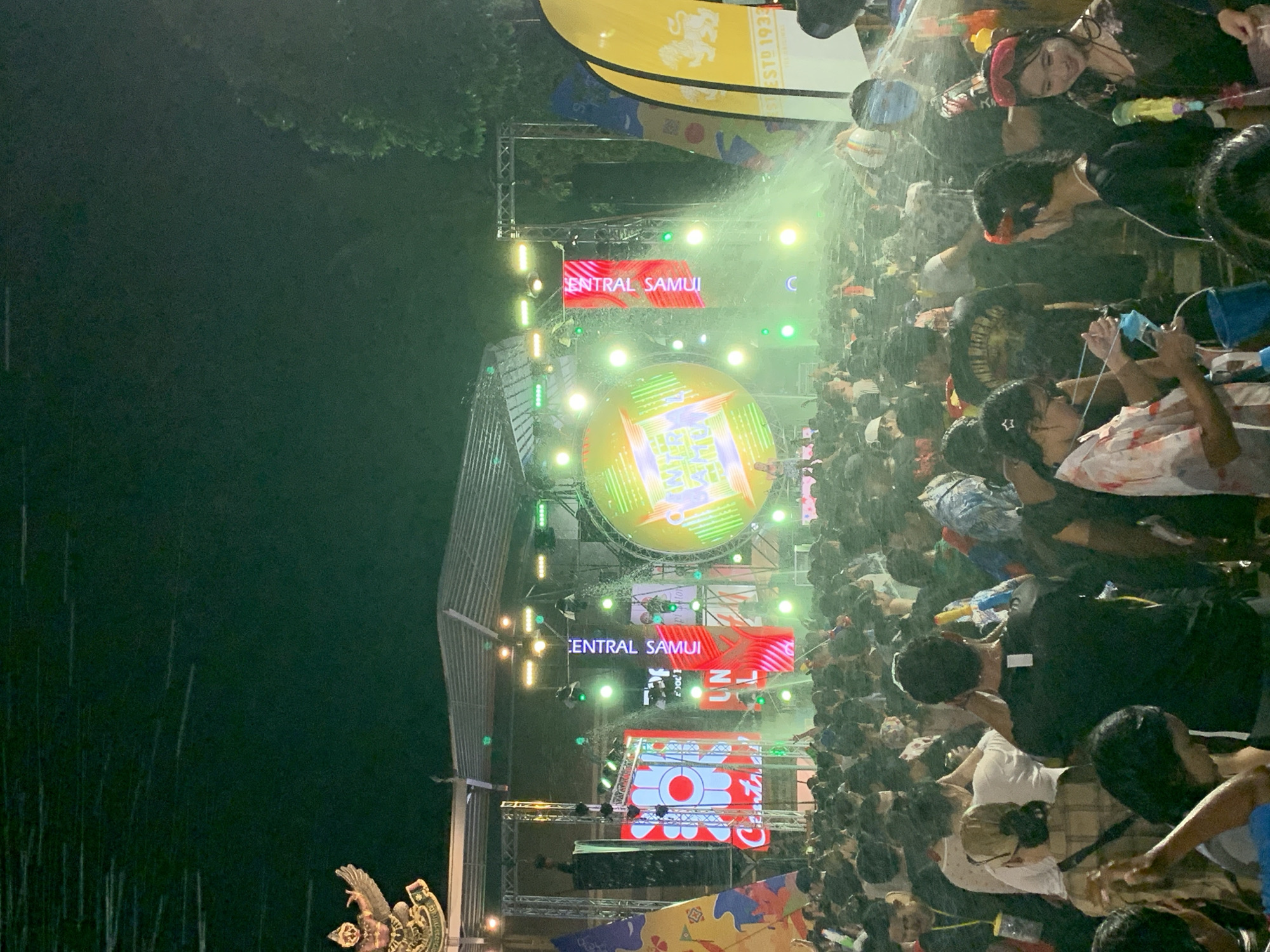 A crowd of people participates in Songkran (Thai New Year), in Baan Chaweng, Thailand. Many people are holding water guns. Water is being sprayed on the crowd from above. Stage lighting and signs are visible above the crowd. The signage says "CENTRAL SAMUI".