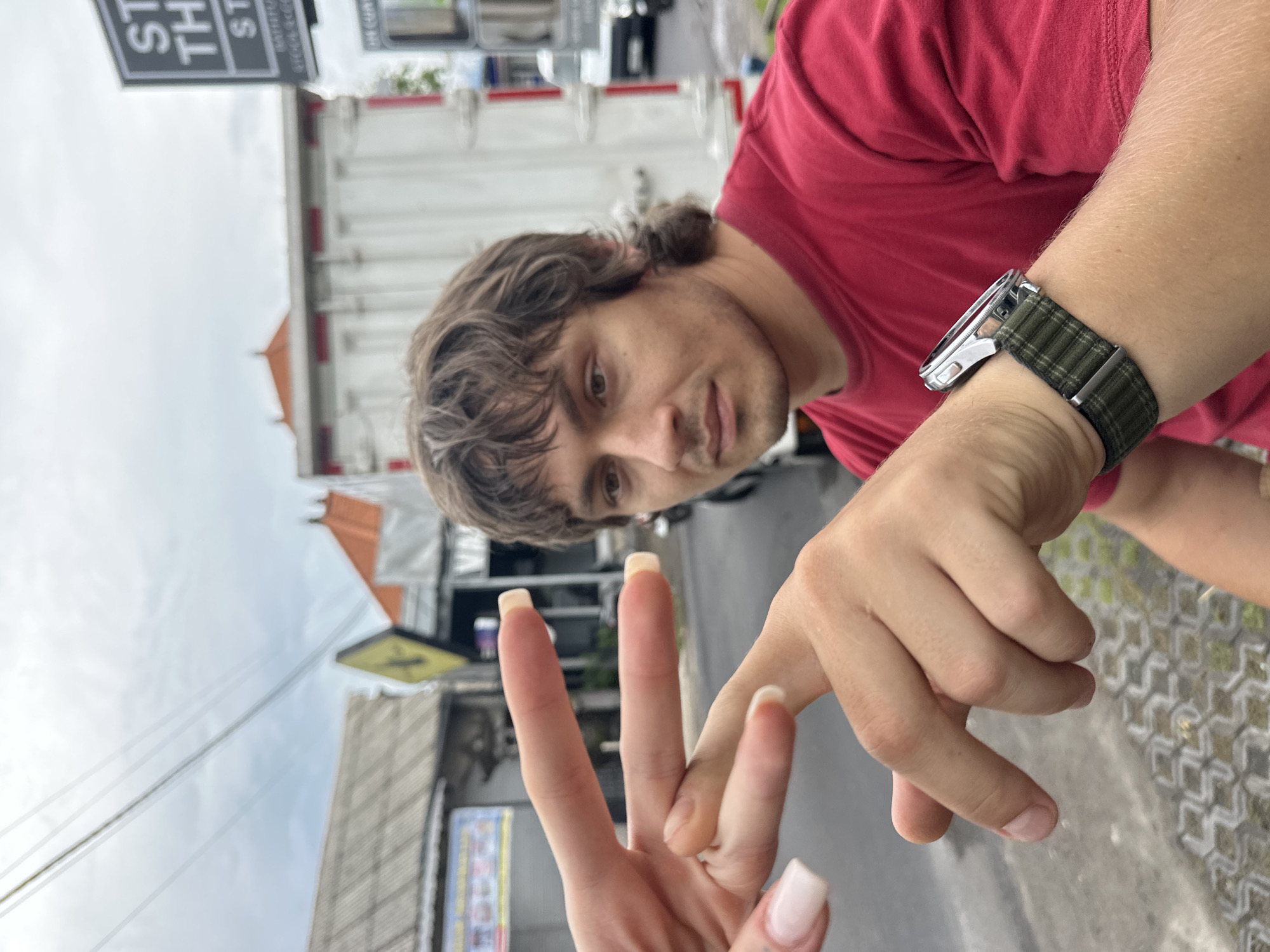 A horizontally oriented, close-up shot depicts a fair-skinned man in a red t-shirt holding his right hand up, which wears a silver watch with a green patterned band on his left wrist. His shoulder is visible on the right. His face is tilted towards the camera. In front of him, a second hand with long, light-pink manicured nails is making a "peace sign." The background indicates an outdoor setting with buildings and street signs visible in Munggu, Indonesia. A white truck with red stripes is partially visible behind the man's head. The sky is overcast.