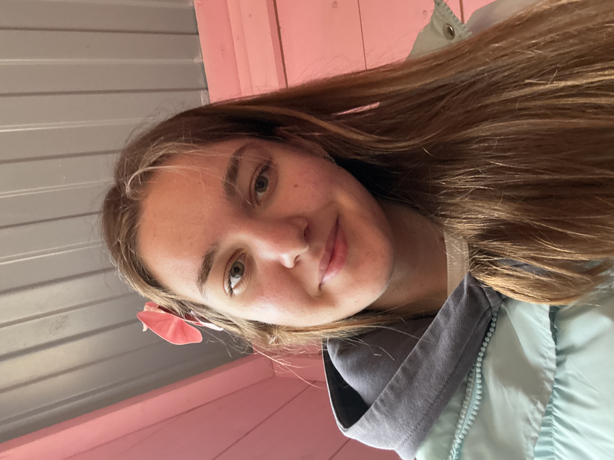 A young woman is captured in a selfie, her head tilted slightly to the left, suggesting an upward angle of the shot. She has light brown hair and a fair complexion. The setting includes a pink and grey colored backdrop, with hints of what might be a building or structure.
It is not possible to determine the location to be Perm, Russia.