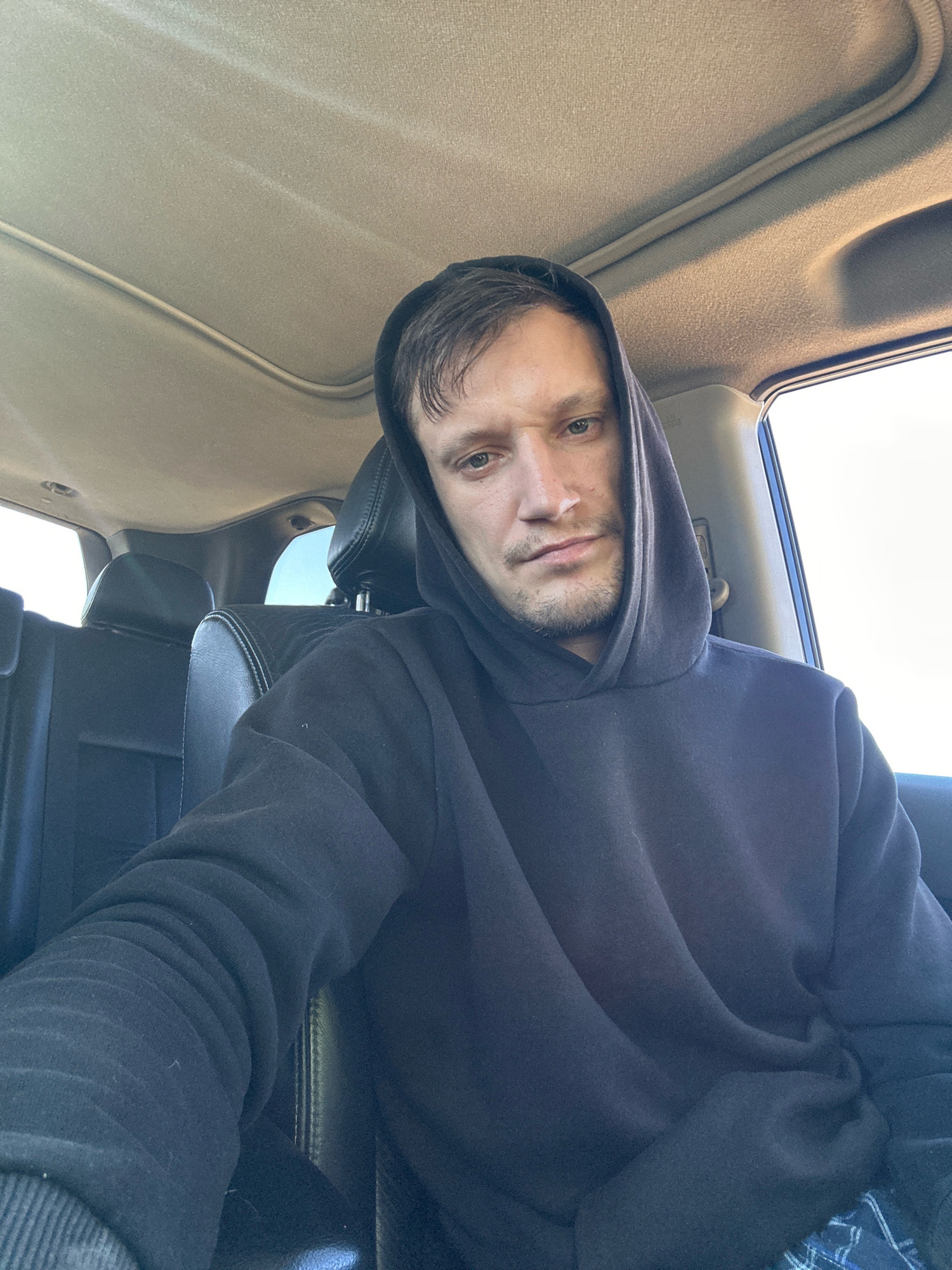 A middle-aged Caucasian man is seated inside a vehicle, looking directly at the camera. He is wearing a dark hooded sweatshirt with the hood up. The interior of the vehicle appears to be that of an SUV, with leather seats visible behind him. Sunlight streams in from the windows and through a sunroof, illuminating the interior. The scene suggests a casual, everyday moment, possibly a selfie taken while traveling. There are no identifiable landmarks or specific details that would indicate the location within Makiv, Ukraine.