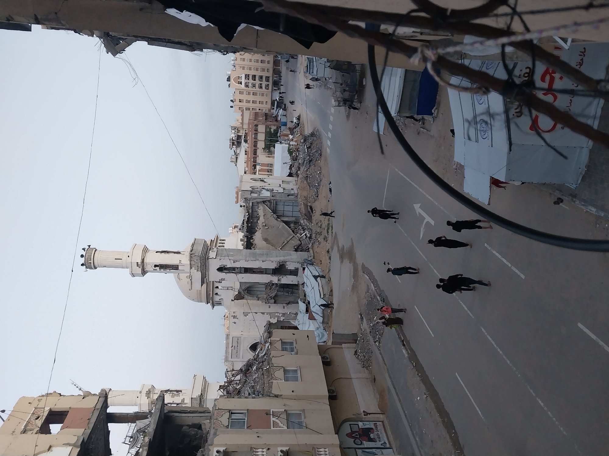 This image depicts a street scene in Khan Yunis, Palestinian Territory, characterized by extensive structural destruction under an overcast sky.
In the background, a severely damaged mosque stands prominently, featuring a white dome and a tall, slender minaret. Large piles of rubble surround its base and extend into the adjacent areas. To the left of the mosque, a multi-story building shows significant damage, with sections of its facade collapsed, revealing interior rooms and a partially collapsed roof. The ground floor of this building displays a sign with "YASEEN GAMFS" text and a red "Squid Game" symbol.
The asphalt road running through the center of the image has visible white lane markings. Several individuals, primarily adult males in dark clothing, walk on the road in the midground. A smaller group, including a woman in a headscarf, walks on the left sidewalk. Further down the street, additional pedestrians are visible. On the right side of the road, beyond the main traffic lane, multiple white, canvas-covered tents, some bearing Arabic script and humanitarian organization logos (e.g., IOM), are erected amidst debris. A smaller structure with a blue roof is also present.
Overhead, multiple black wires crisscross the frame, suspended between damaged buildings. The immediate foreground, suggesting the viewpoint from an elevated and damaged structure, includes dark structural elements and wires. Buildings in the distant background also show varying degrees of damage.