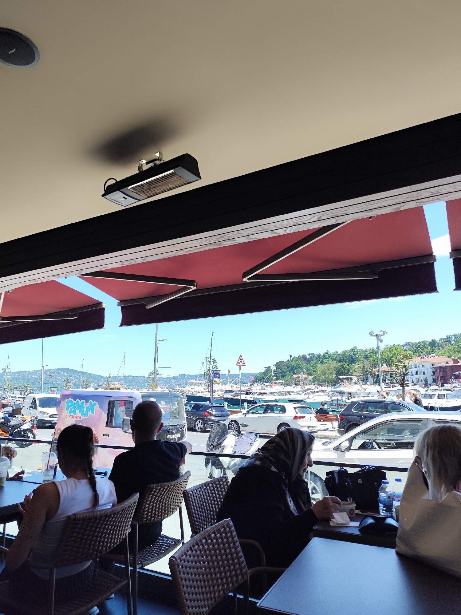 The image shows people sitting at tables under an awning, likely at a restaurant or cafe in Sarıyer, Turkey, as indicated by the presence of local brands (such as Pamuk Çikolata) and the view of a marina with numerous boats and a cityscape. The scene is captured during daylight, with cars and other vehicles visible in the background, suggesting a lively outdoor setting in Sarıyer.