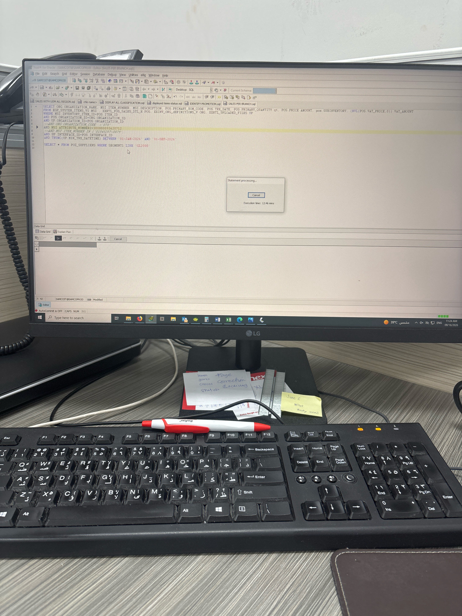 The image shows a desk with a computer monitor, keyboard, and some sticky notes. The monitor displays a SQL query execution window with a progress bar and an execution time of 12:46 minutes. The SQL query seems to be for retrieving sales data. The keyboard is a standard QWERTY layout with some Arabic characters. The sticky notes have handwritten text, some of which appears to be in English and Arabic. The overall environment appears to be an office setting. There is no visible indication of the location being Dhahran, Saudi Arabia.