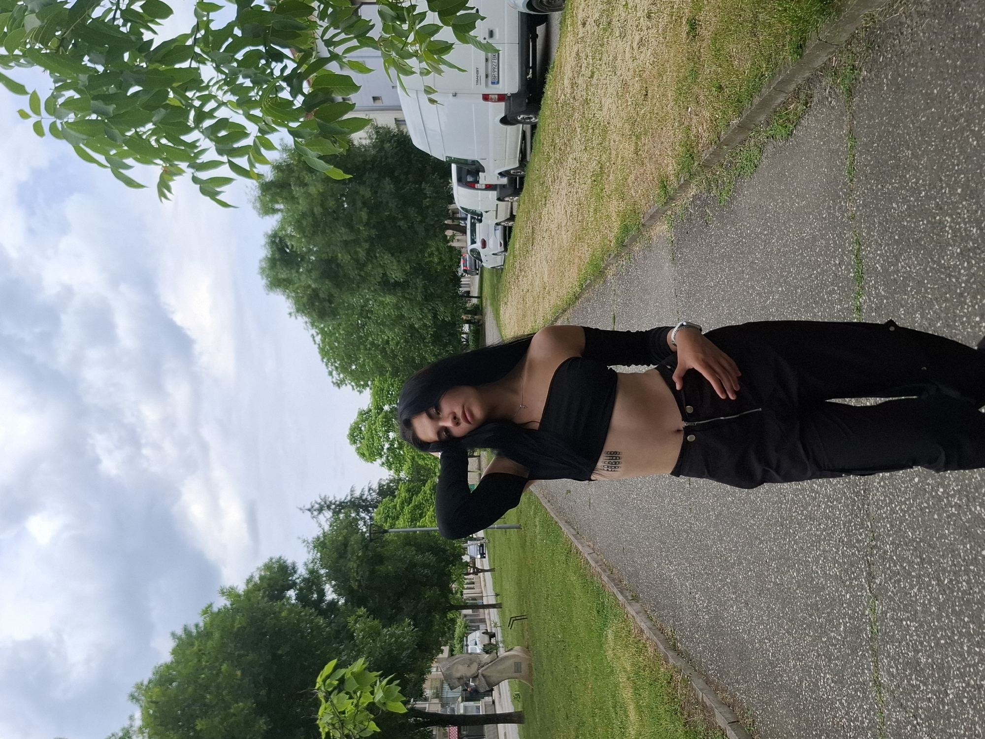 A woman with long black hair, wearing a black midriff-baring top and black pants, stands on a paved pathway in what appears to be a city park. She rests her head on her left hand. There are trees and green grass to her left and right. Behind her, further down the pathway, are white vans and cars parked. The sky is cloudy. The location is likely near the city of Mezdra, Bulgaria, due to the presence of a vehicle with a license plate beginning with the letters "BP".