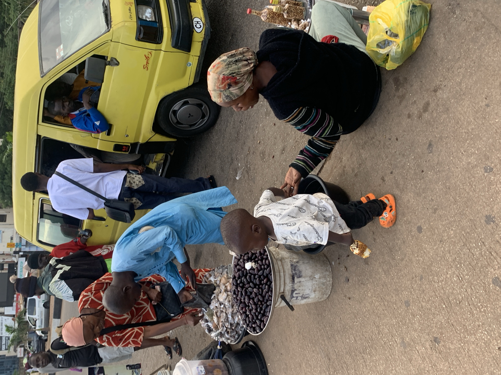 A group of people are gathered around a yellow commercial van, possibly a "danfo" bus, on a paved street, likely in Jos, Nigeria. In the foreground, a woman wearing a headscarf and a patterned top is sitting next to a young child dressed in a white t-shirt and orange Crocs. The child is holding a piece of food. Next to them, a man in a blue kaftan is interacting with a woman in a red and white patterned dress who is standing near a large tray of dark, round fruits, possibly olives or plums. Behind them, several other people are visible, including a man driving the yellow van. The environment appears to be a busy urban street with a few cars and buildings in the background. The overall scene depicts a common street market or transportation hub in a Nigerian city.