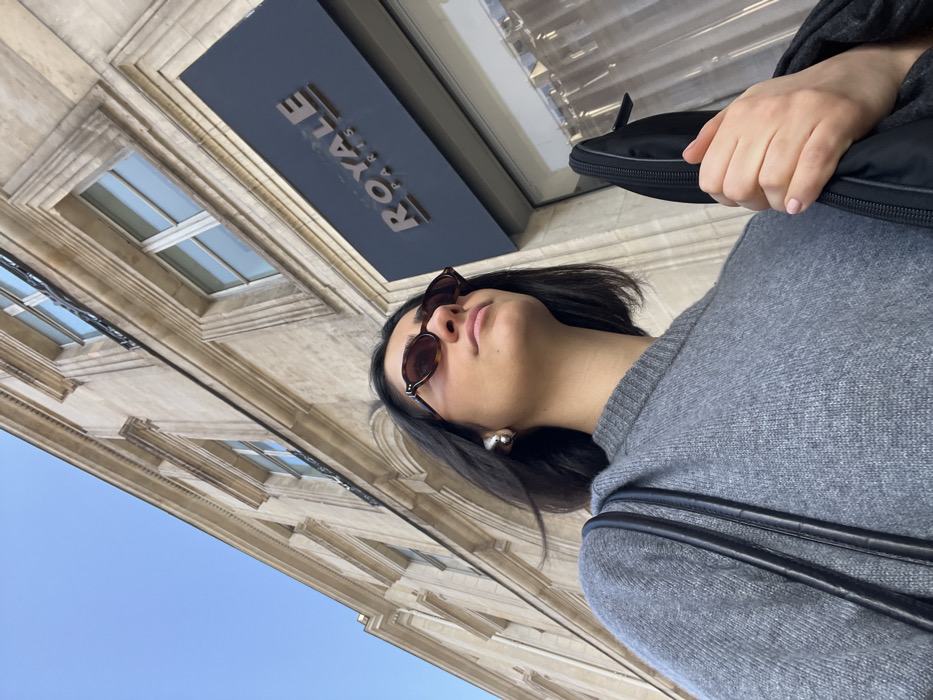 A low-angle shot captures a female subject positioned outdoors in Paris, France, against a classical stone building. The woman, with dark, short hair, wears oval, tortoiseshell-patterned sunglasses, a grey crew-neck knitted sweater, and a silver-toned, abstract-shaped earring on her left ear. A black strap, possibly from a bag, is visible over her left shoulder. Her right hand, with light pink manicured fingernails, holds a black zippered case. She gazes upward and to the right. The background features a multi-story, light-colored stone building characteristic of Parisian architecture, displaying ornate window frames and rectangular windows. A prominent black rectangular sign on the building's facade reads "ROYALE PARIS" in raised metallic lettering. A window next to the sign is obscured by a translucent curtain. A clear blue sky occupies the upper portion of the frame. The presence of the "ROYALE PARIS" sign suggests the location is near Rue Royale or within the city of Paris.