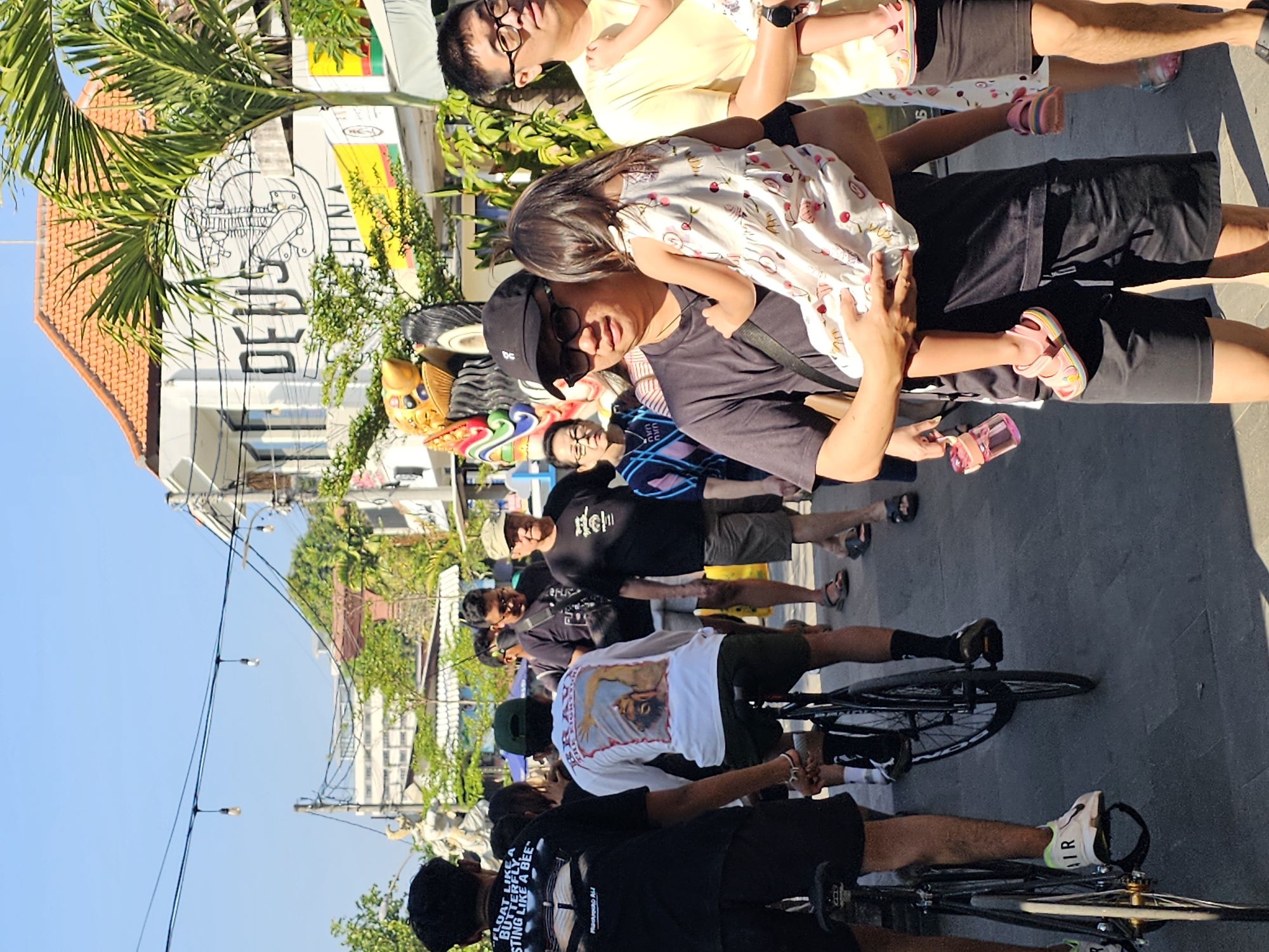 In Denpasar, Indonesia, a street scene unfolds under a clear blue sky. The foreground is dominated by a man carrying a young girl, both dressed in casual attire. The man wears a black t-shirt, sunglasses, and a black baseball cap, while the girl is in a patterned dress and pink sandals. To their right, another man in a yellow t-shirt and glasses holds another child. Behind them, a group of people are walking, including a man in a dark t-shirt and shorts, and an older man in a light-colored cap and t-shirt. Two individuals on bicycles are also visible, their legs and bikes taking up the lower portion of the frame. The backdrop features a building with the "DEUS" logo prominently displayed, along with palm trees and other foliage, suggesting a commercial or tourist area. The street is paved, and string lights hang across the scene, illuminated by the bright sunlight.