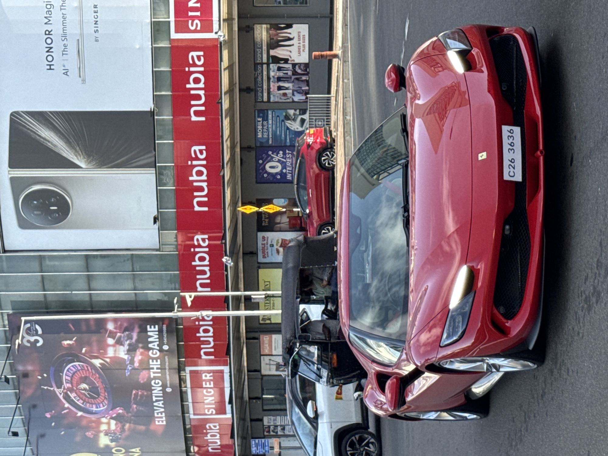 The image shows a red Ferrari with license plate C26 3636 on a street in Colombo, Sri Lanka. Multiple cars and a tuk-tuk are nearby. Billboards display advertisements for Honor Magic, a casino, Nubia phones and Singer appliances. Several commercial buildings are visible. Pedestrians are visible in the background.