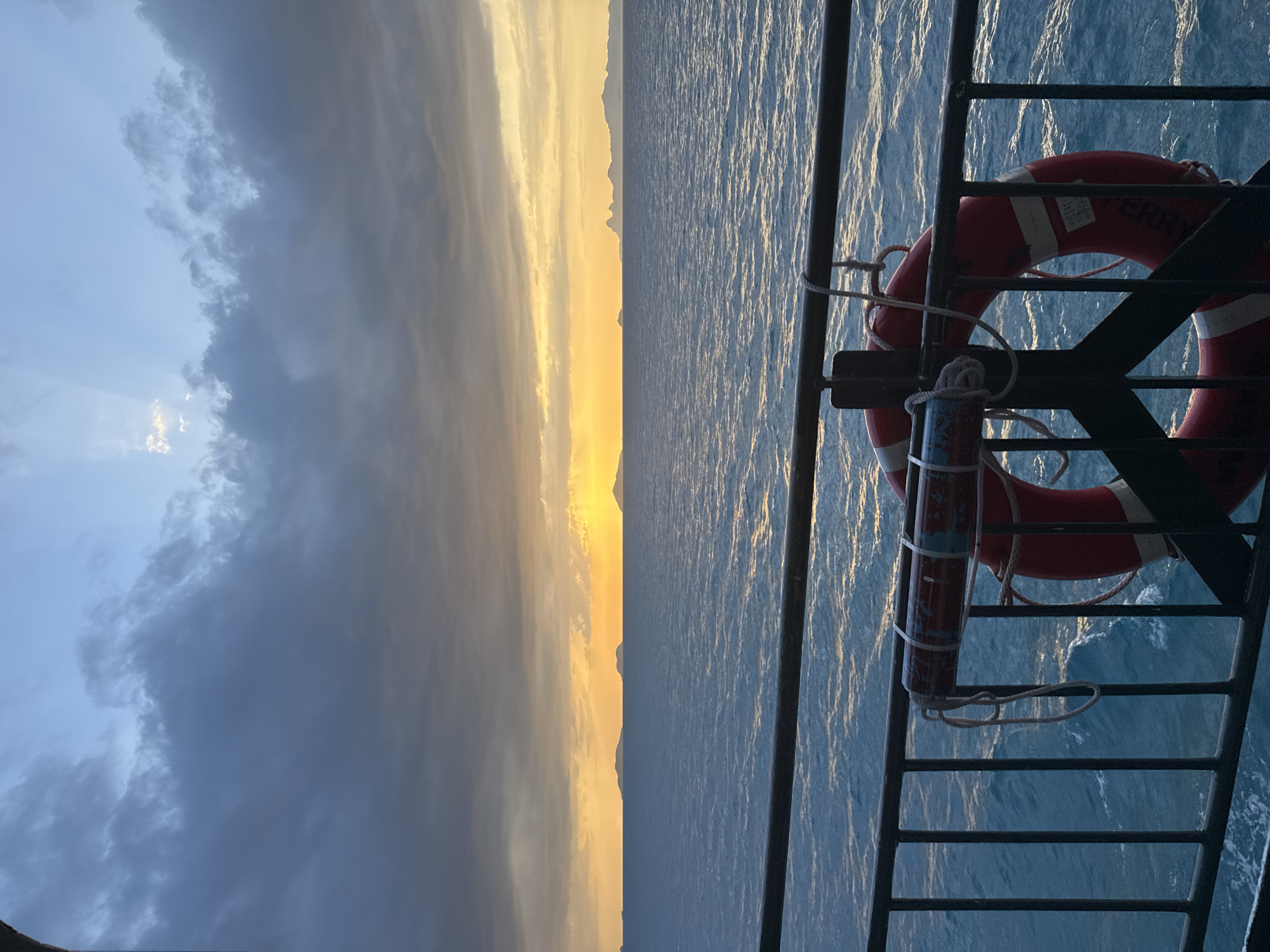 A sunset over the ocean is visible from a ferry in Thailand. The sun is low on the horizon, casting a warm orange glow across the sky and water. Islands can be seen in the distance. A life preserver hangs on the railing of the ferry in the foreground. The water is a deep blue, with small waves creating a textured surface. The sky is a mix of blue and grey clouds, with some rays of sunlight breaking through.