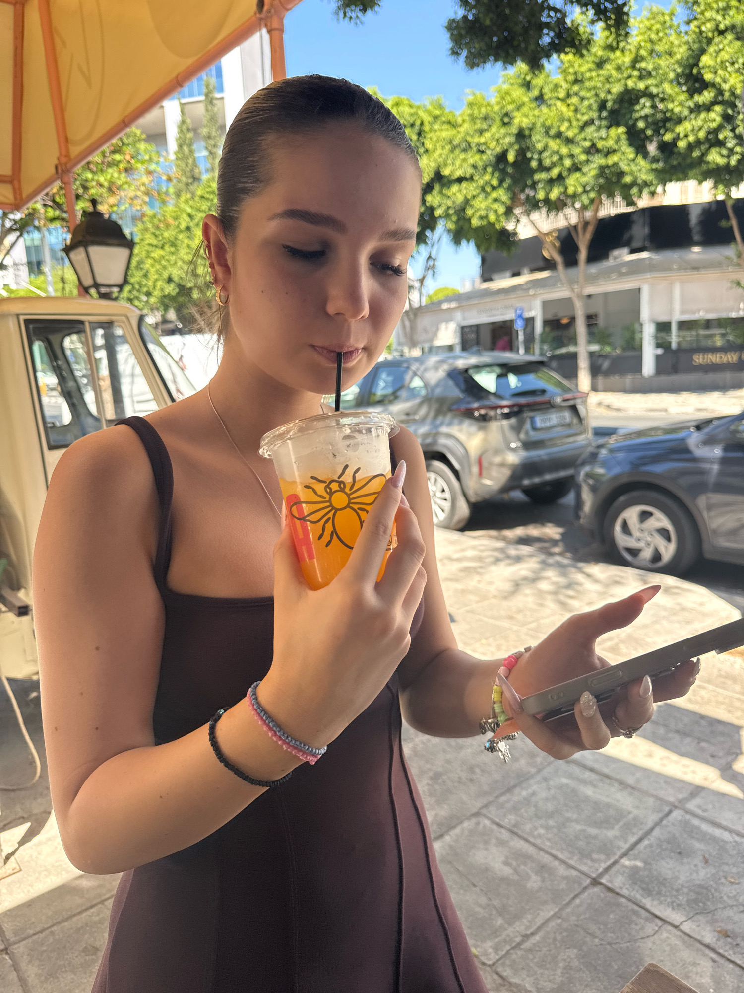 A young woman with her hair pulled back is standing outside, holding a plastic cup with an orange beverage and a straw. She is looking down at her phone, which she is holding in her right hand. She is wearing a dark-colored dress and has bracelets on her left wrist and rings on her fingers. In the background, there are trees, buildings, and cars, suggesting an urban environment. The overall impression is a casual outdoor setting, possibly a cafe or street. The image appears to be taken in Limassol, Cyprus, based on the contextual clues of the urban landscape and vegetation.