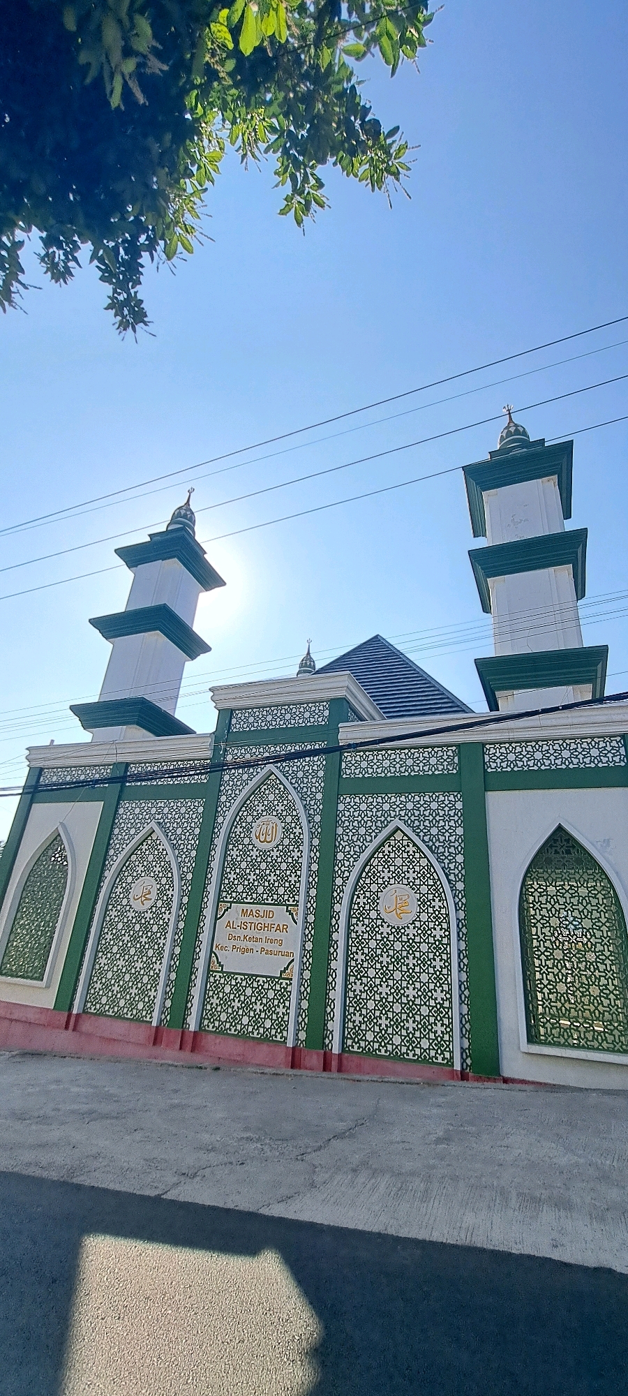 The image shows a mosque in Ketanireng, Indonesia, under a clear blue sky. The mosque has a white and green facade with intricate geometric patterns. Two minarets with green tops rise above the main building. The name of the mosque, "MASJID AL-ISTIGHFAR," is visible on a plaque on the front wall, along with the location details: "Dsn. Ketan Ireng, Kec. Prigen, Pasuruan." Green foliage from a tree hangs into the top left corner of the frame. Power lines are visible in the sky. The ground in front of the mosque appears to be a road or paved area, with a shadow cast across it.