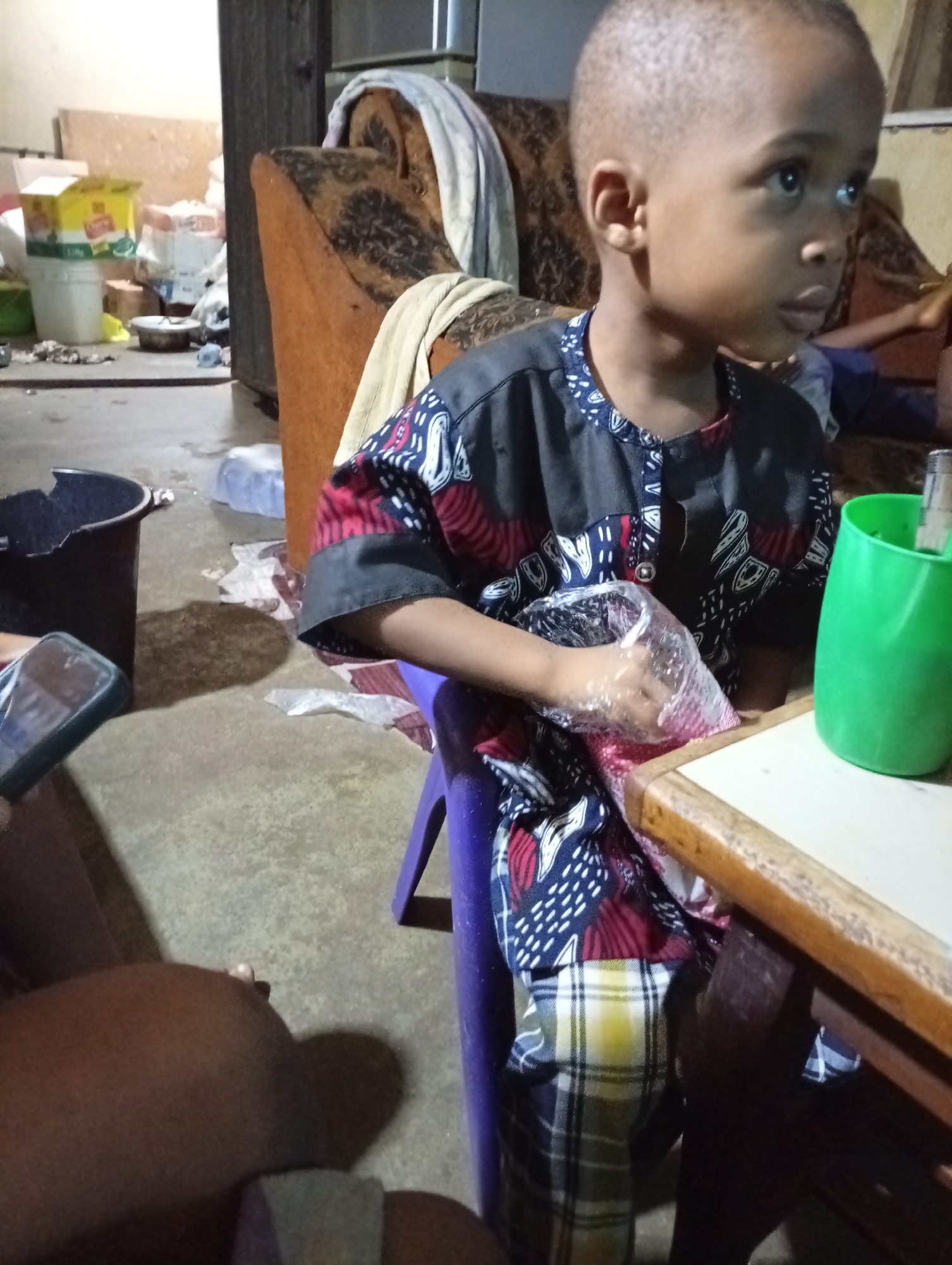 A young boy is sitting at a wooden table indoors. He is wearing a colorful patterned shirt and plaid pants. He has a clear plastic bag in his hands. On the table is a green plastic cup. The boy is looking to his right. An adult's leg and hand holding a smartphone are visible in the lower left corner. The floor and background are cluttered. A brown patterned sofa is behind the boy. The location is Unknown, Unknown.