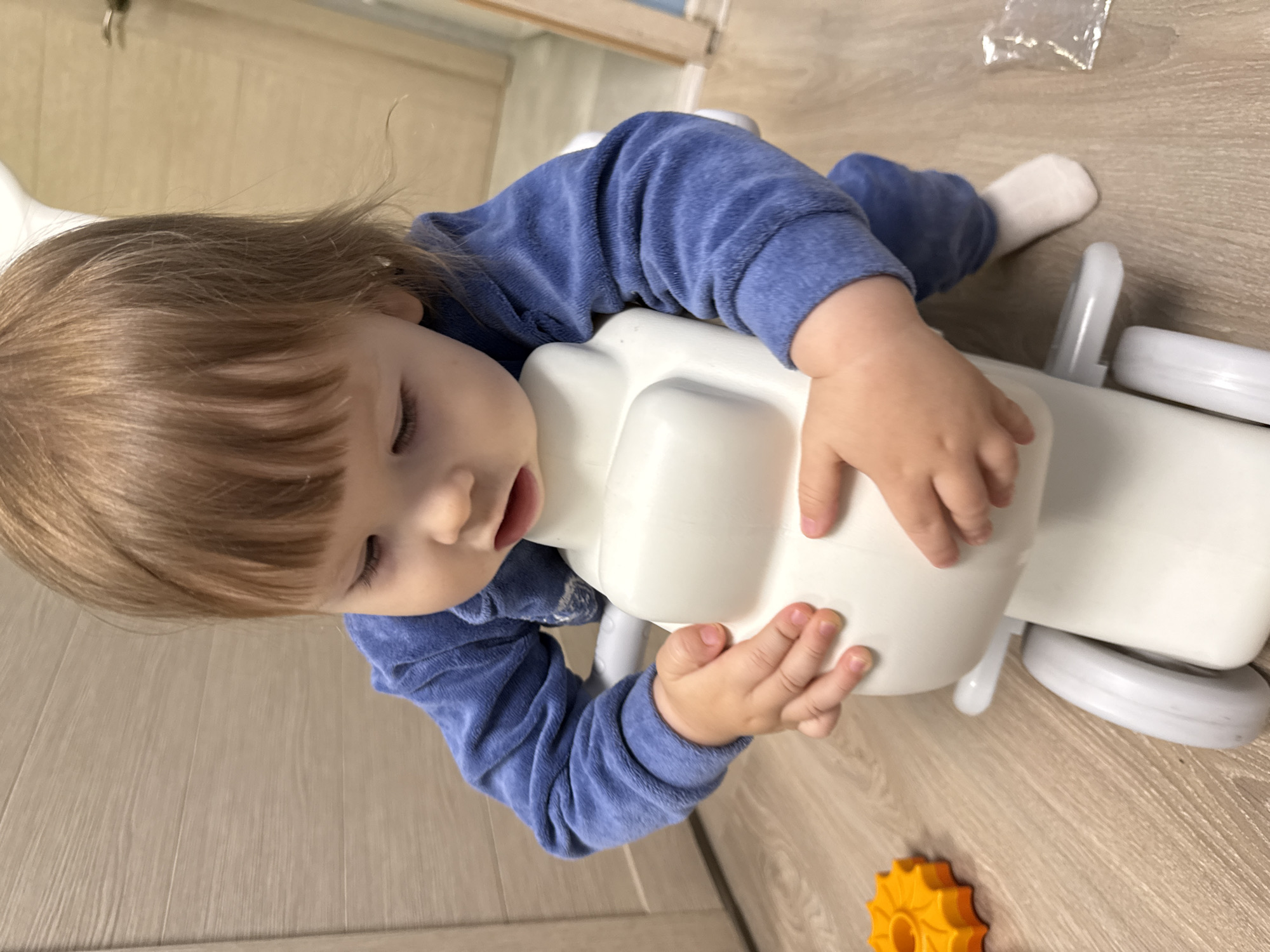 A toddler, wearing a blue velour tracksuit and white socks, is playing with a white toy motorcycle on a light brown wooden floor. The child's hands are gripping the handlebars of the toy. To the left of the child, an orange toy gear is visible. The background appears to be a domestic interior with a wooden door or wall paneling. There is no indication that this scene is located in Moscow, Russia.
