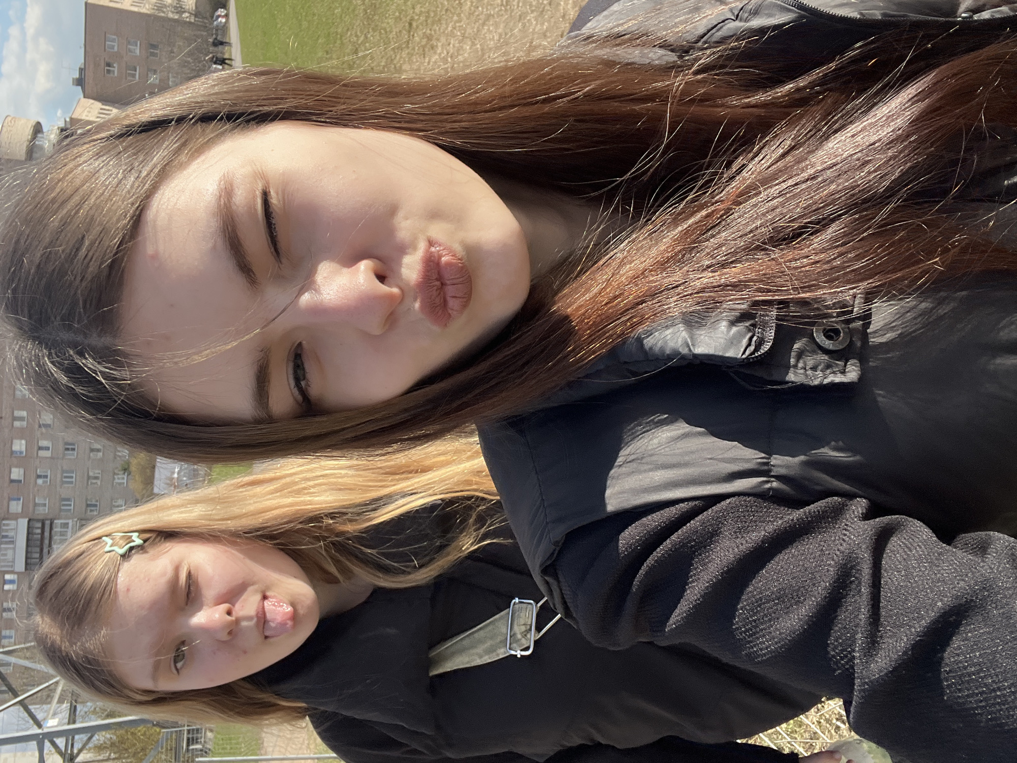 Two young women are captured in a selfie. The woman in the foreground has brown hair and is making a kissing face with her eyes mostly closed. She is wearing a black jacket. Behind her, a second woman with blonde hair is sticking out her tongue and winking. She also wears dark clothing. The background includes an apartment building, a green field, and a partial view of a historical-looking tower. Given the architectural style, the scene is likely located in Saint Petersburg, Russia.