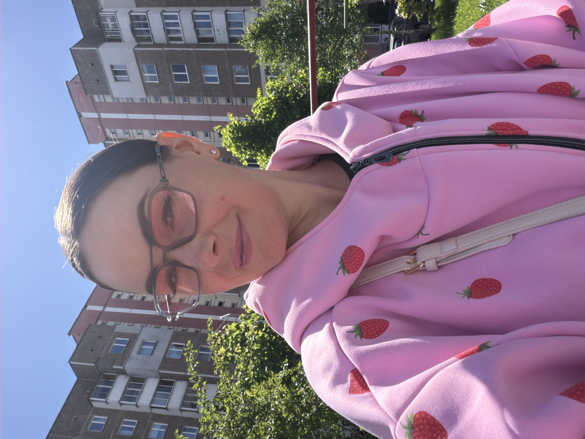 A woman with fair skin is pictured in front of a building and trees in an urban setting. She is wearing spectacles with rose-tinted lenses. The hoodie is pink with a repeating strawberry print and is zippered. A cream-colored purse strap is visible across her chest. The building behind her is multi-story and features a brick and grey facade with regular window placement. Green trees partially obscure the building and are likely street trees. The sky is blue. The location appears to be Unknown, Unknown.