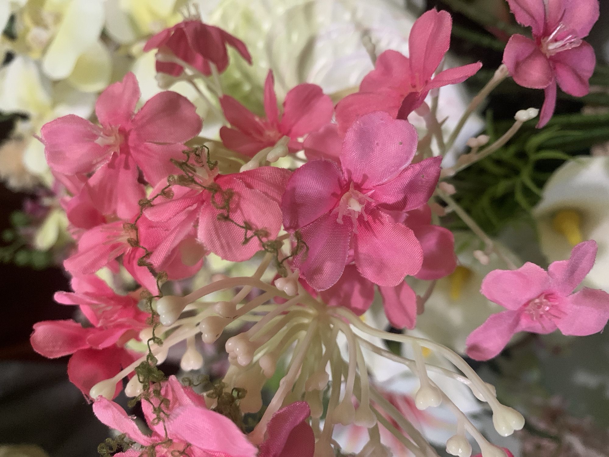 The image shows a close-up view of a floral arrangement featuring numerous bright pink flowers with delicate white stamen. The flowers appear to be artificial, with a fabric-like texture visible on their petals. Interspersed among the pink blossoms are small, pale green buds or accents, and some thin, brown, dried elements that resemble tendrils. The background is a soft blur of lighter colors, possibly other flowers or foliage, creating a shallow depth of field that emphasizes the vibrant pink blooms in the foreground. The lighting is soft and diffused, highlighting the textures and colors of the flowers without harsh shadows. The location of this floral arrangement in Kuala Pembuang, Indonesia, is not discernible from the visual information provided in the image.