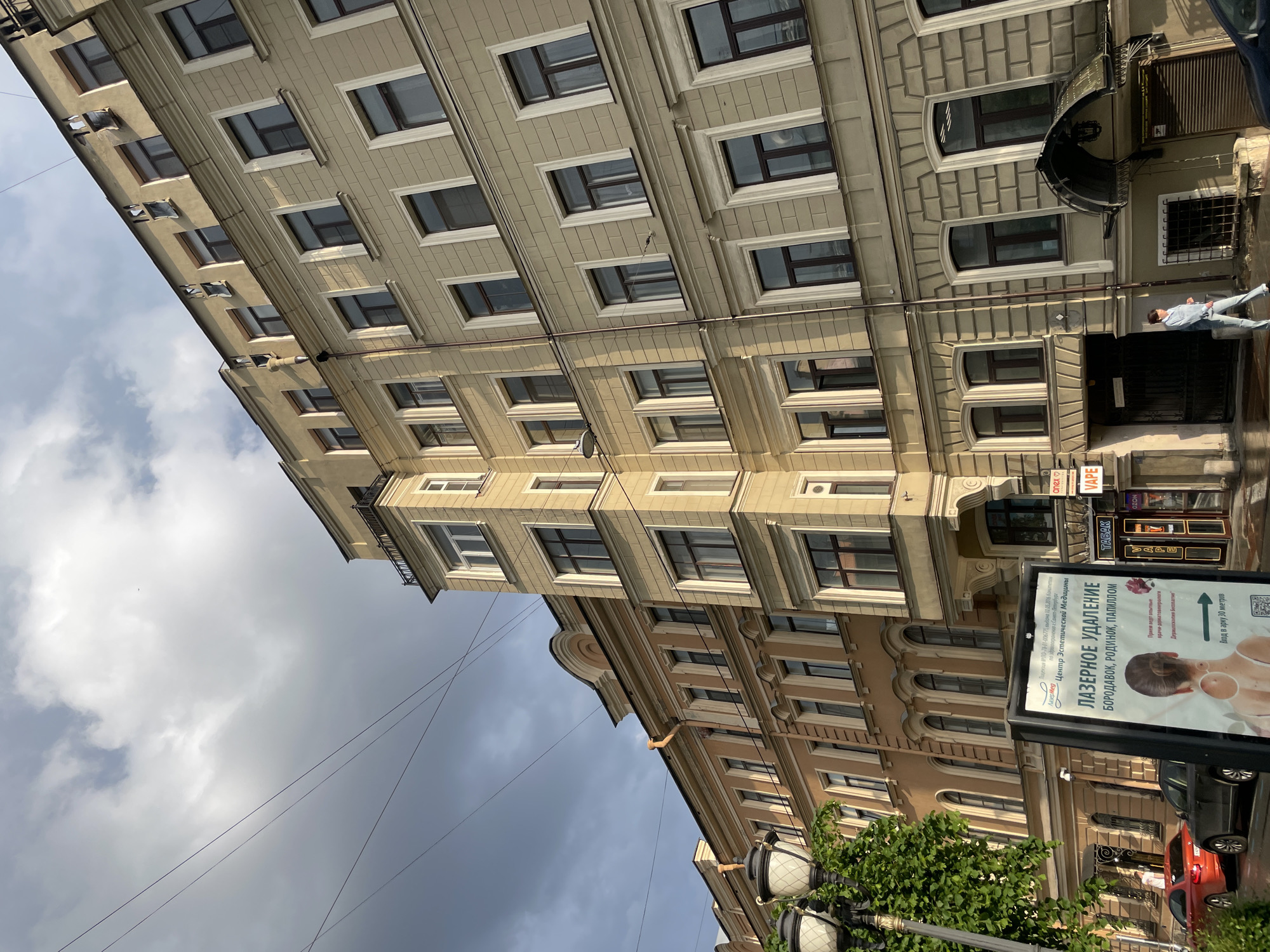 The original image, tilted counter-clockwise, depicts a vibrant street scene, likely in Saint Petersburg, Russia, based on the visible Cyrillic text and provided context.
A tall, light beige-colored building with numerous rectangular windows, many featuring white frames, dominates the left side of the frame. Several small air conditioning units protrude from its upper floors. Overhead wires crisscross the partly cloudy sky above.
On the right, another multi-story building stands. Its facade is a slightly darker, warm beige with elaborate architectural details, including rusticated stonework on the lower floors and decorative cornices. The ground and first floors feature arched windows and doorways, with subsequent floors displaying rectangular windows, some with small balconies or ledges. An ornate, curved iron balcony or awning is prominent above an entrance on the first floor. At street level, various commercial signs are visible, including "VAPE", "ТАБАК" (Tobacco), and "OZON". A person in light-colored jeans and a top stands near an entrance on the sidewalk.
In the foreground on the right, a large, rectangular street billboard prominently displays an advertisement for "ЛАЗЕРНОЕ УДАЛЕНИЕ БОРОДАВОК, РОДИНОК, ПАПИЛЛОМ" (Laser Removal of Warts, Moles, Papillomas) from "Центр Эстетической Медицины" (Center of Aesthetic Medicine) / "Лазер Мед" (Laser Med). The billboard includes a graphic of a person's neck and shoulder, a QR code, and details such as "Лицензия... Комитету по здравоохранению г. Санкт-Петербурга" (License... Committee for Healthcare of Saint Petersburg) and "Вход в арку 30 метров" (Entrance through the arch 30 meters).
A decorative black streetlight, partially obscured by green foliage, is seen in the bottom right. Parts of two cars, including a red one, are visible parked on the street. The sky is partly cloudy with patches of blue. The scene appears to be set during daytime, with bright ambient light and soft shadows suggesting late morning or early afternoon.