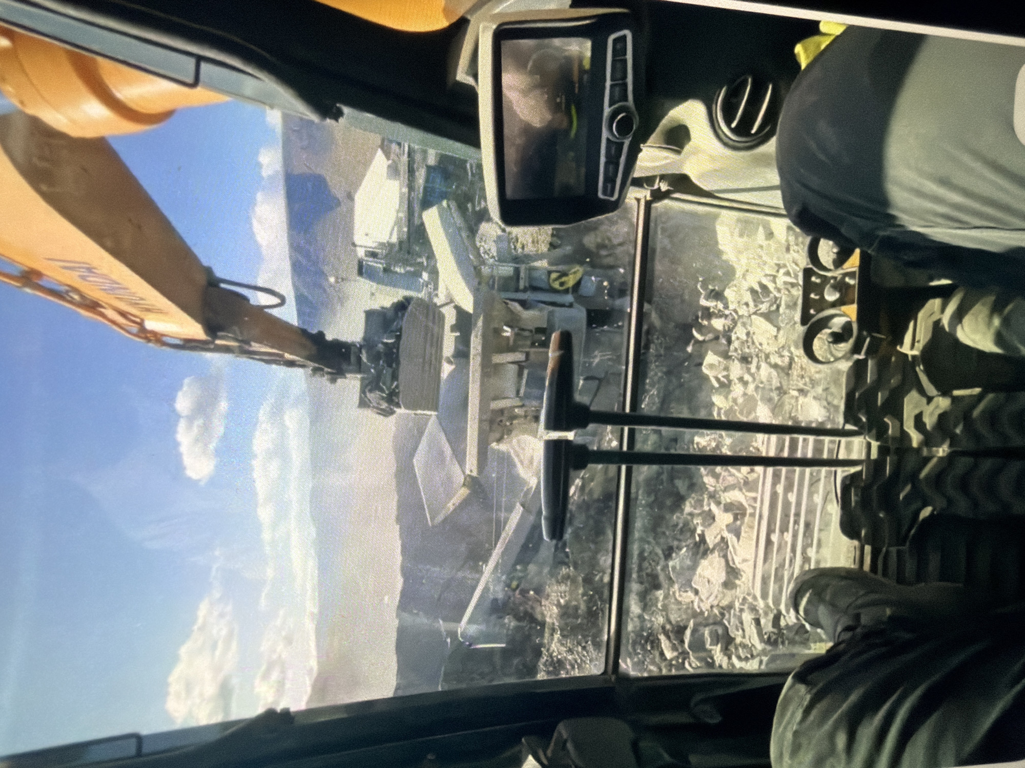The image is taken from the interior of a construction vehicle, likely an excavator, looking out at a demolition site. In the foreground, the operator's legs are visible, clad in dark pants. Two control levers are positioned centrally. To the right, a digital screen displays a blurry image, possibly a rear-view camera feed.
The exterior view reveals a scene of destruction. A large excavator arm, emblazoned with the "HYUNDAI" logo, is actively engaged in moving debris. Below it, a collapsed structure, composed of concrete and metal, is being dismantled. The ground is covered in rubble and rocks, suggesting a significant demolition operation. In the background, rugged hills or mountains are visible under a bright blue sky with scattered clouds. The overall environment appears to be an outdoor construction or demolition zone.