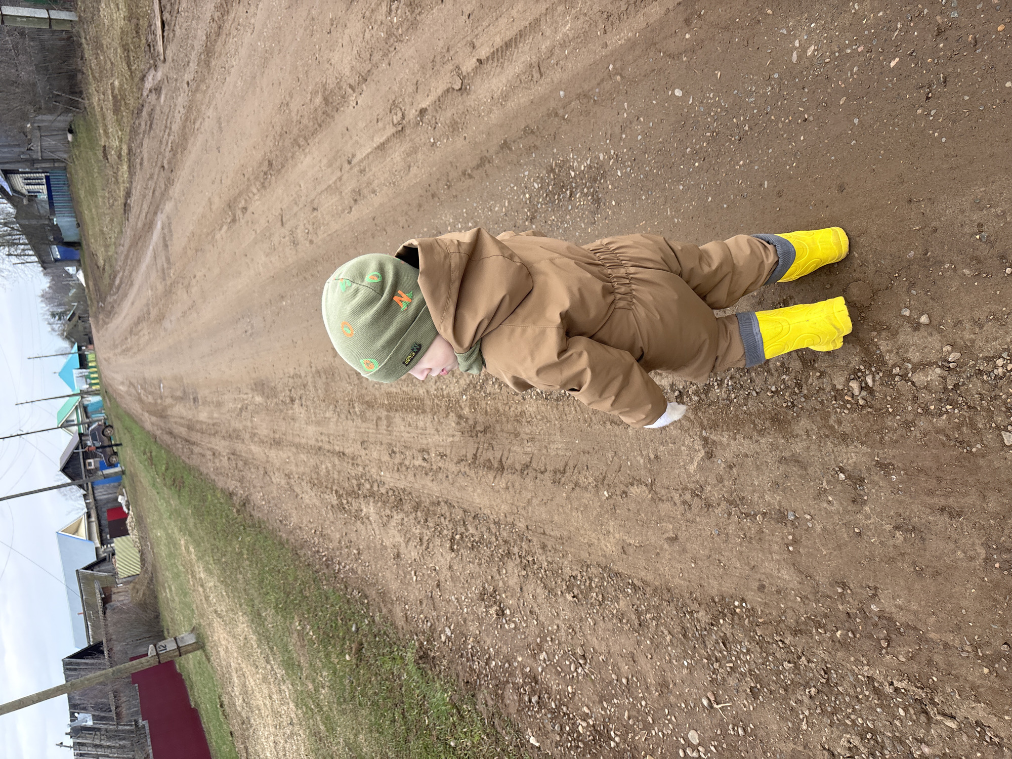 The original image is oriented sideways, depicting a small child lying face down on a dirt road. The child, likely a toddler, is dressed in a full-body brown or khaki-colored suit and bright yellow rubber boots. A light green beanie with orange and green patterns, including the letters 'N' and 'O', covers the child's head. The child's arms are extended forward on the ground, suggesting an activity like crawling or playing.
The setting is an unpaved, wide dirt road with visible tire tracks and scattered pebbles. To the left of the road, a strip of sparse grass separates it from a series of rural houses or structures, featuring various colors and roof styles. Bare trees and utility poles are also visible along the roadside, indicating a non-urban environment, possibly a village or countryside. The sky appears overcast, providing soft, diffused light, and the ground shows signs of dampness, hinting at recent rain or snowmelt. The time of day is daytime. No other specific text is clearly legible beyond the letters on the beanie.