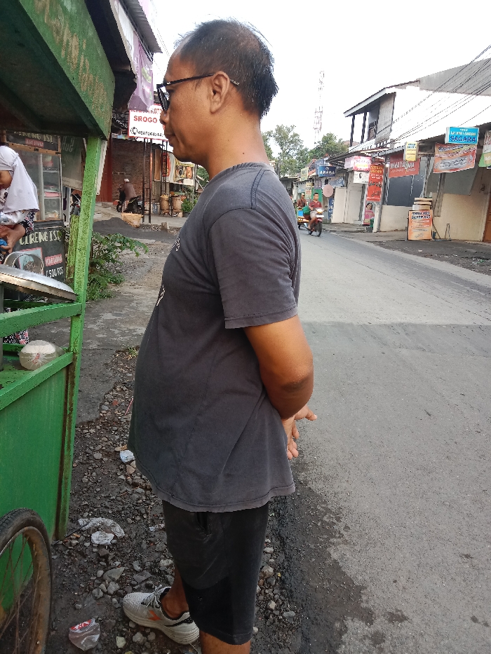 An individual, identified as a male of apparent Southeast Asian ethnicity, stands with his right side facing the viewer on a gravelly shoulder adjacent to an asphalt road in Sukomulyo, Indonesia. He wears a dark grey short-sleeved t-shirt, black shorts, and dark-framed eyeglasses, with his hands clasped behind his back. His gaze is directed towards the left. To his immediate left, a green, wheeled street food cart is partially visible, where a second individual wearing a light-colored head covering is positioned, leaning into the cart. The cart displays Indonesian text, including "CIRENG ISI" and "500 PCS," along with a white styrofoam container. The ground beneath the standing man consists of loose dirt, small rocks, and scattered debris, including a discarded plastic bottle. In the mid-ground, the asphalt road extends into the distance, flanked by a row of single and two-story buildings of simple construction on the right side. These buildings feature various commercial signs in Indonesian. Further down the street, multiple individuals and at least two motorcycles are discernible. A tall communications tower is visible in the distant background against a pale sky.