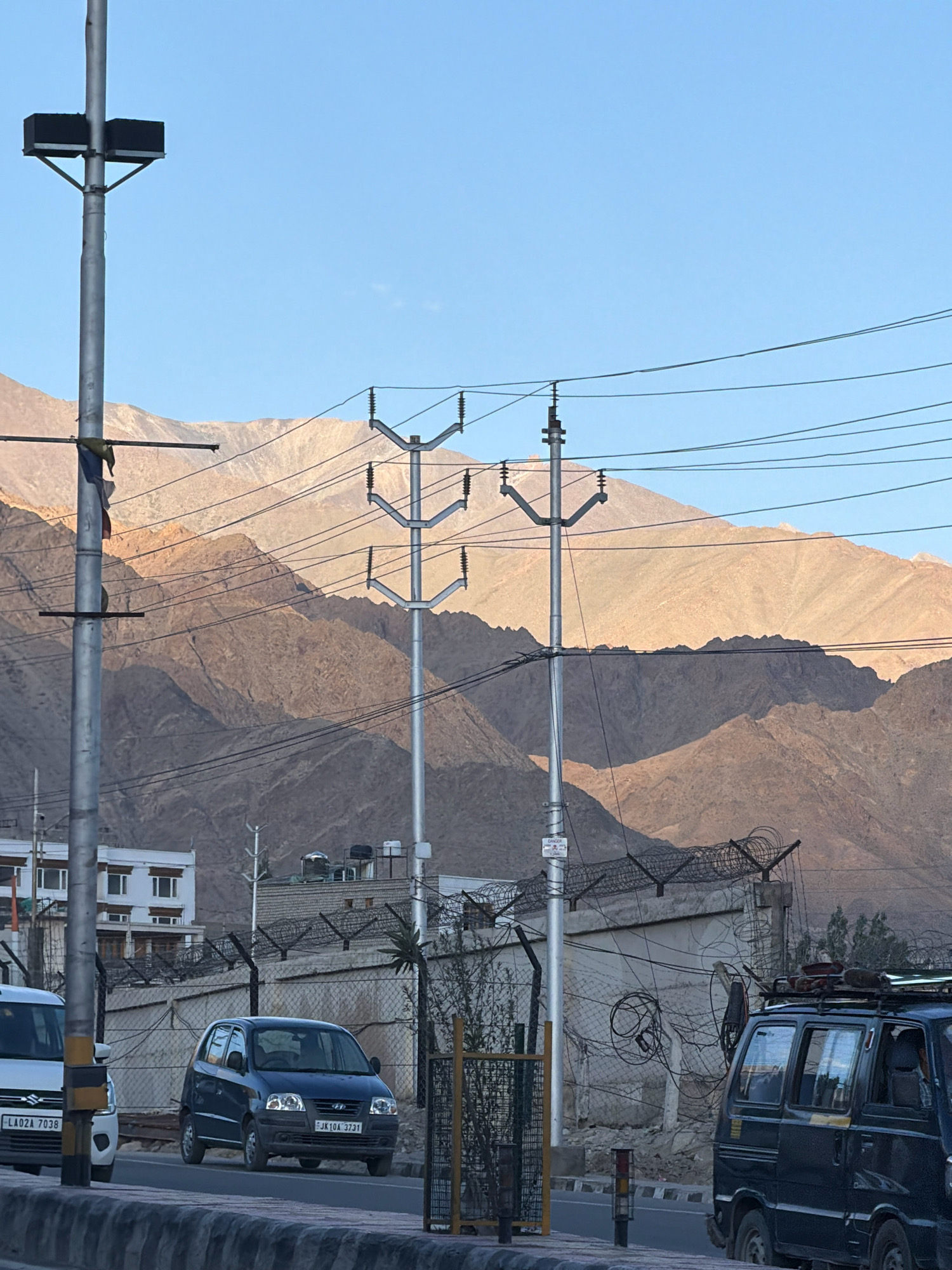 The image depicts a street scene in Leh, India, with a clear blue sky and rugged, sun-drenched mountains in the background. A prominent utility pole with two black boxes, likely lights or communication equipment, stands on the left. Multiple power lines crisscross the frame, supported by utility poles. In the mid-ground, a concrete building with barbed wire fencing is visible. Two vehicles are on the road: a dark blue hatchback car with license plate "JK 10A 3731" in the center, and a dark blue van with a roof rack on the right. A white vehicle with license plate "LA02A 7038" is partially visible on the far left. The road is bordered by a stone embankment and a metal fence. The overall environment suggests an arid, mountainous region.
