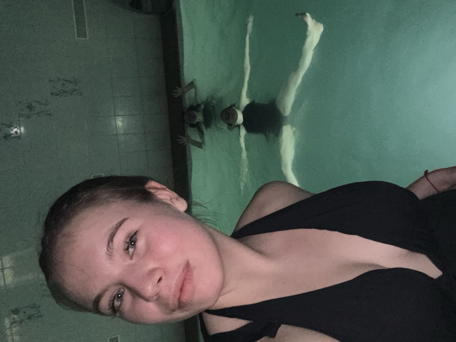 A selfie of a woman in a black top with a pool in the background. Another woman is on the pool's edge with her hands on the wall. A third person floats on their back in a crucifix position in the pool. The pool and the tiled wall give a greenish cast to the image.