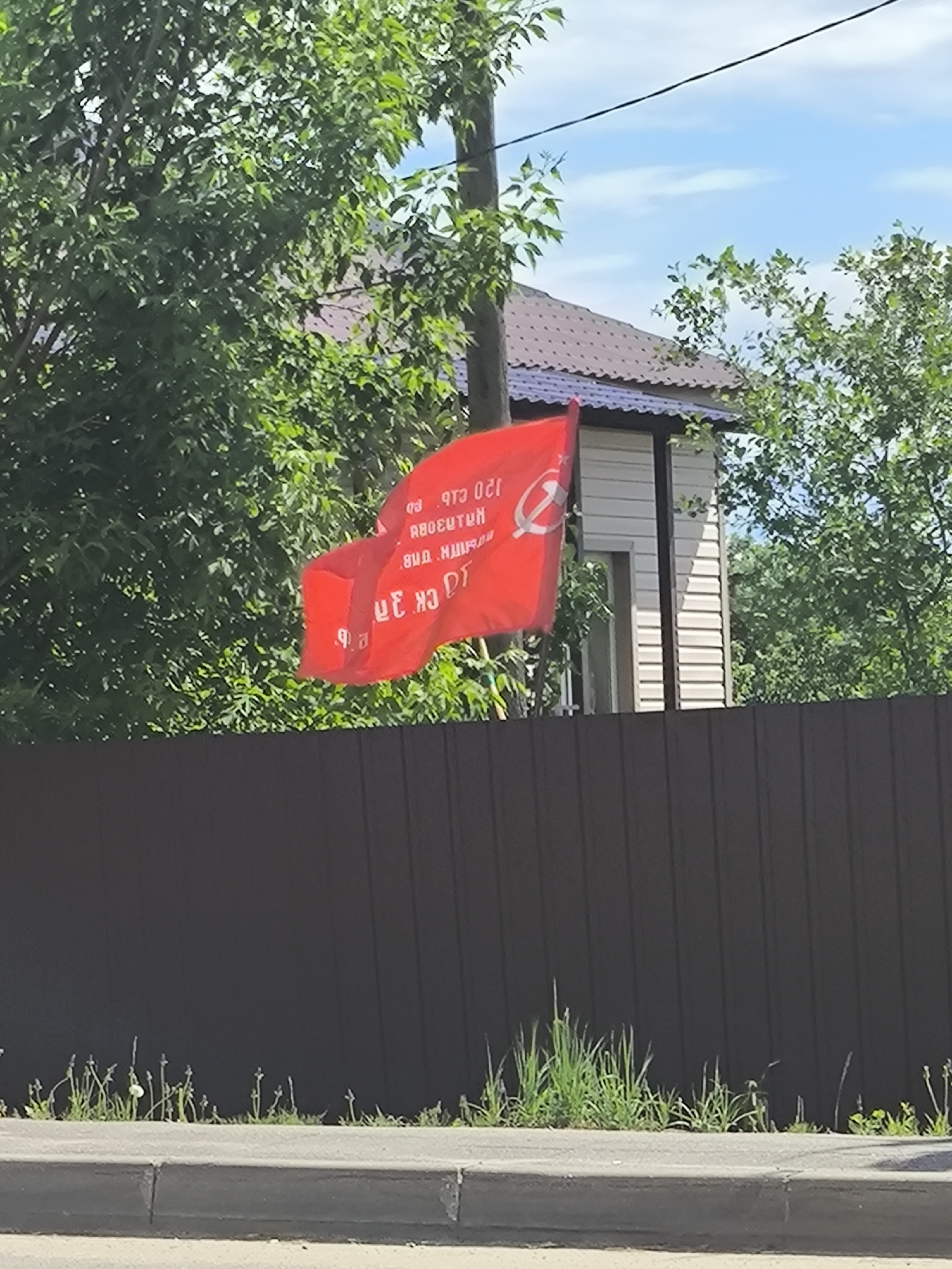 A red flag with an emblem resembling a hammer and sickle hangs on a pole behind a brown fence in an outdoor, daytime setting. Text is printed on the flag in a language other than English. Behind the flag and fence, there are trees, a house with light-colored siding, and a brown roof. Grass and weeds grow along the sidewalk in front of the fence.