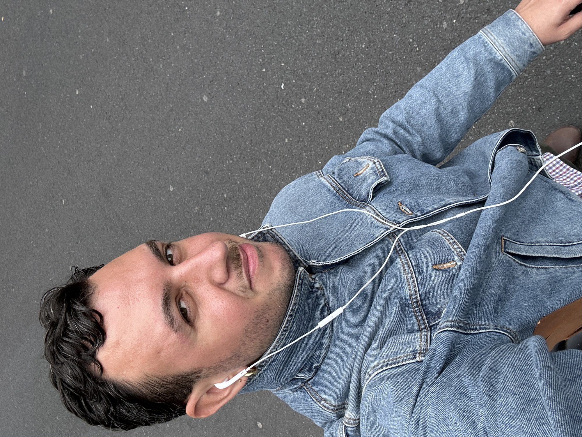 A man is wearing a denim jacket and white earphones. The backdrop is grey asphalt. The setting may be in London, United Kingdom.
