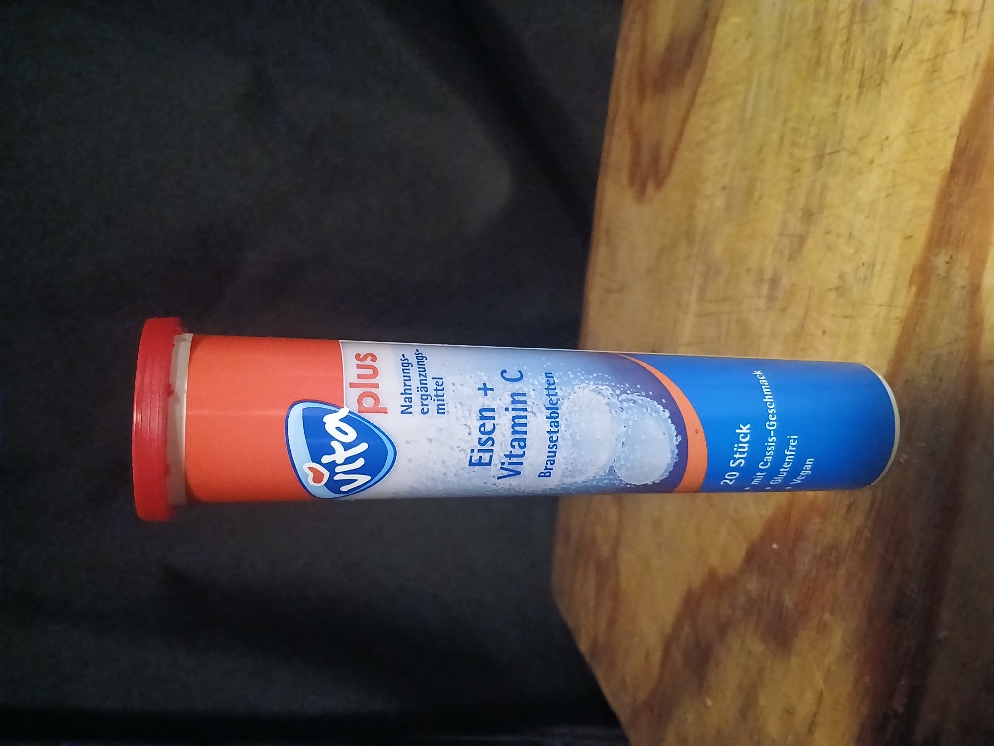 The image displays a tube of Vita + Eisen + Vitamin C effervescent tablets. The tube is primarily orange at the top, transitioning to white with blue text and graphics lower down. The brand "Vita" is prominently featured in a blue logo with a heart. Text on the tube indicates it is a dietary supplement and specifies "Eisen + Vitamin C Brausetabletten" (Iron + Vitamin C Effervescent Tablets). The bottom portion of the tube shows details such as "20 Stück" (20 pieces), "mit Cassis-Geschmack" (with blackcurrant flavor), "Glutenfrei" (gluten-free), and "Vegan". Effervescent tablets are visible through the lower part of the translucent tube, submerged in a blue liquid with bubbles. The tube is standing upright on a wooden surface, which appears to be a table or countertop. The background is dark and out of focus. There are no people or specific city landmarks visible that would indicate a location in Douai, France.