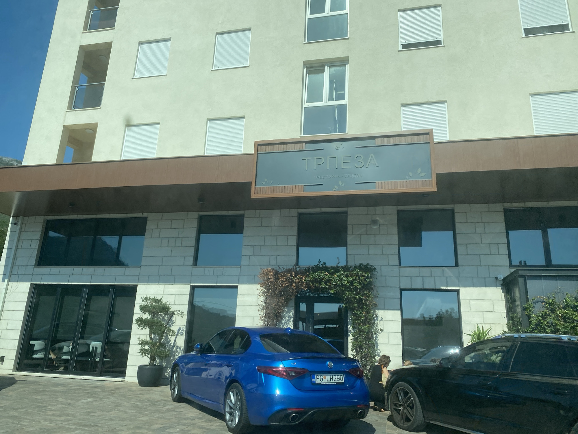 The image depicts the facade of a building, likely a hotel or restaurant, in Sutomore, Montenegro. The building features a light-colored exterior with multiple windows, some of which are covered by rolling shutters. A prominent sign above the entrance reads "ТРПЕЗА" in large Cyrillic letters, with smaller text below that appears to be "PESTO PAН - TRPEZA". The sign is framed by a wooden border and accented with leaf motifs.
In front of the building, a bright blue Alfa Romeo sedan is parked, facing away from the viewer. The license plate is visible and reads "PG LH290". To the right of the blue car, a portion of a black SUV is visible. A woman with brown hair is standing next to the rear of the black SUV, facing the building. Another person, partially obscured, is also near the black SUV. The ground in front of the building is paved with a patterned material. Lush green vines are growing on the building's facade, particularly around the entrance area. To the left of the entrance, a potted plant with green foliage is situated. The sky is clear and blue.