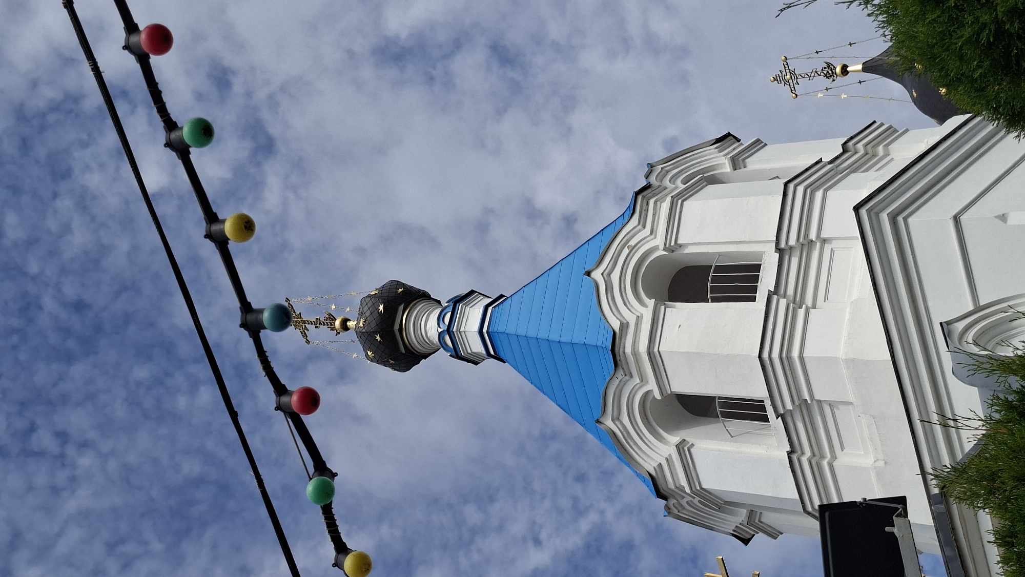 The image captures a low-angle view of a white Orthodox church dome with a blue roof, topped with a black cupola adorned with gold stars and a golden cross. The structure is partially obscured by green foliage in the foreground. Above the church, a string of colorful lights, red, yellow, and green, is strung across a dark wire against a blue sky with white clouds. The scene appears to be set in Pastavy, Belarus, judging by the architectural style and the context of the location. There are no people or discernible actions in the frame. The lighting suggests it is daytime.