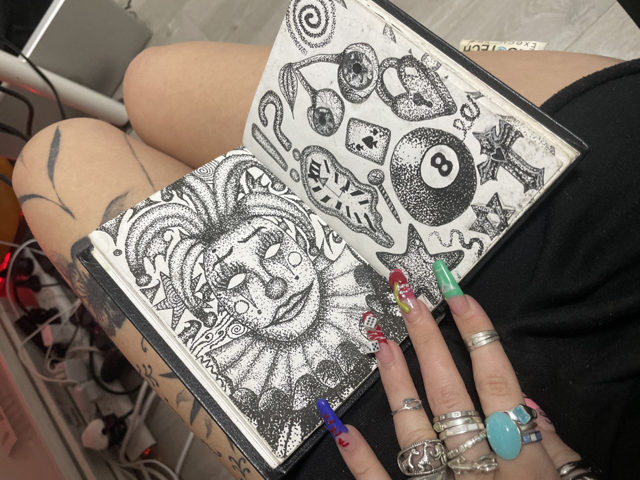 An open sketchbook displays detailed pen drawings: one page features a clown face with intricate dot work, while the opposite shows various symbols like an 8-ball, question mark, hourglass, and cross. The sketchbook rests on a person's lap, whose hand, adorned with multiple silver rings and colorful, elaborately decorated nails, partially covers the drawings. A visible tattoo decorates the person's thigh, adding to the image's artistic and alternative aesthetic.