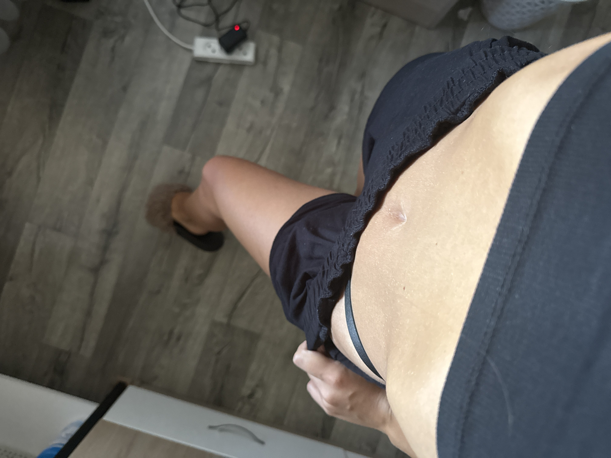 The image shows a person's midsection from a high-angle perspective, focusing on their abdomen and upper thighs. The person is wearing a black sports bra and black shorts that have been pulled down slightly, revealing a portion of a black thong. A small, light-colored scar is visible on the person's abdomen. The floor is made of grey wood-look laminate. In the background, a white electrical outlet with a red-lit plug is visible on the floor. To the right, a white surface, possibly furniture, is partially visible. The image does not contain any information that would allow for the determination of its location in Basarabeasca, Moldova.