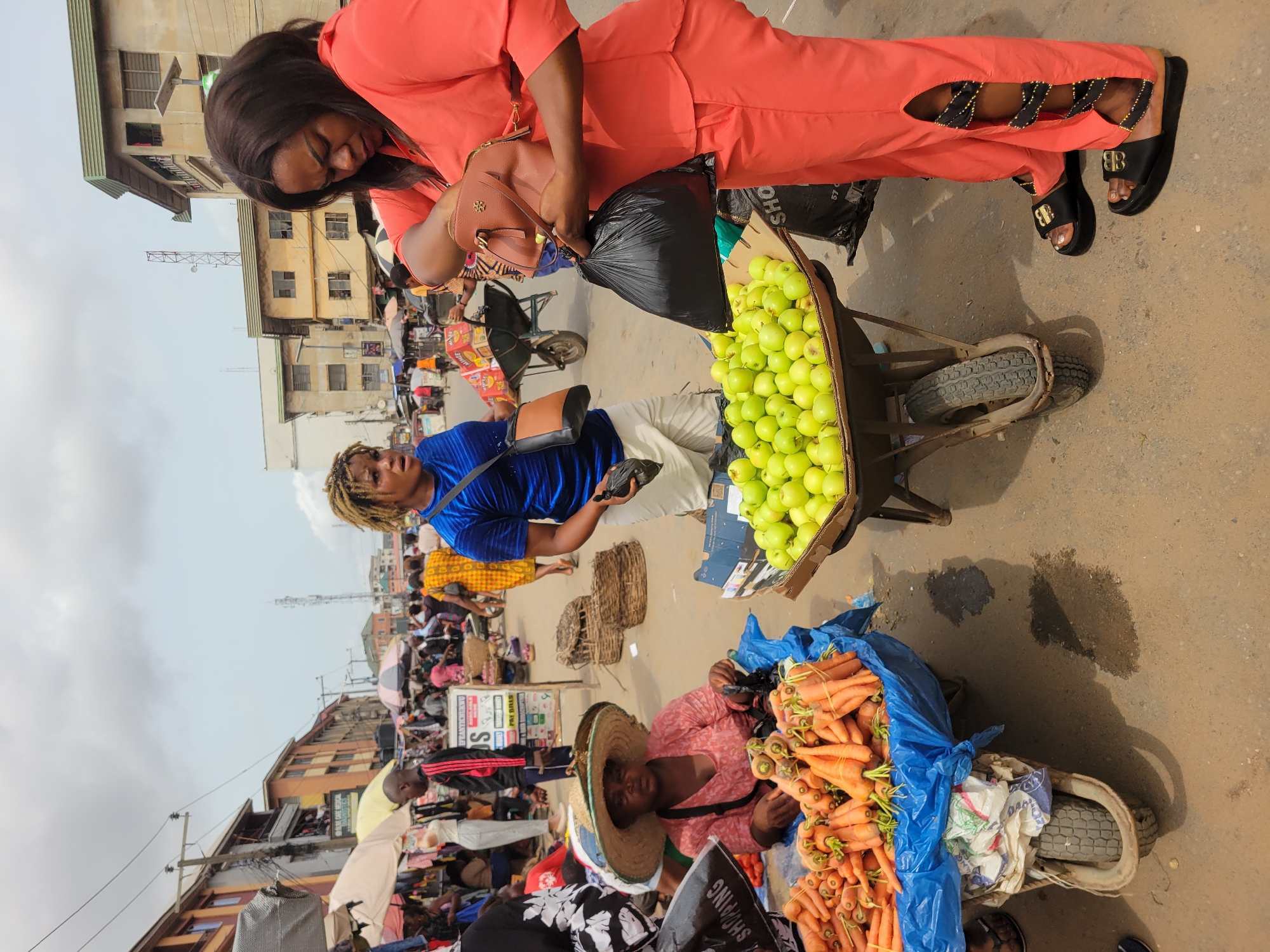 This image captures a vibrant and bustling street market scene in Onitsha, Nigeria, under an overcast daytime sky. The perspective is slightly angled, showing the action from a slightly elevated viewpoint looking down into the market.
In the foreground, a woman wearing a coral-colored top and trousers with distinctive black and yellow patterned cut-outs on the lower legs stands, holding a brown leather purse (with a "Tory Burch" logo) and a black plastic shopping bag. Her black sandals feature a "BB" logo. Adjacent to her is a wooden wheelbarrow overflowing with numerous bright green apples, likely for sale.
To her left, a man with blonde dreadlocks, dressed in a blue velvet-like shirt and light-colored pants, is looking down at his phone. Further into the scene, closer to the ground, a woman wearing a wide-brimmed straw hat and a pink top is seated. In front of her, a large quantity of fresh carrots is arranged within a blue tarp, which appears to be placed on a motorcycle or moped. She seems to be a vendor attending to her wares.
The background is filled with the activity of a busy market. Numerous other people are visible, some standing, some seated, amidst various stalls and merchandise. Several umbrellas are scattered throughout, providing shade. Buildings, some with multi-story windows and weathered facades, line the street, indicating an urban setting. Visible text includes "SHOPPING" on a black bag, "SHOES" partly visible on another bag, and "SALES" on a distant sign. The overall scene depicts a lively commercial hub centered around the trade of fresh produce and other goods.
