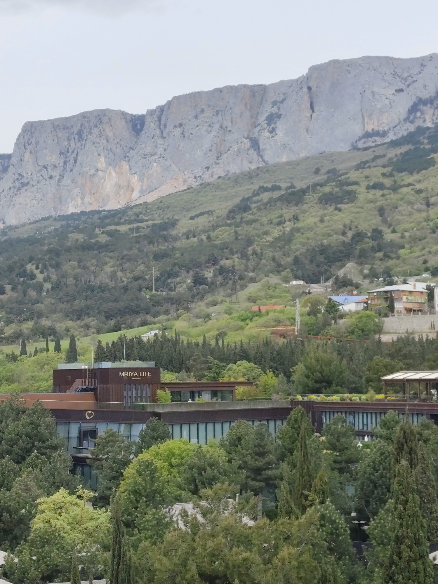 This image presents a panoramic view of a mountainous landscape, likely situated in the Симеизский поселковый совет region of Ukraine. The background is dominated by a majestic, rugged mountain range, characterized by steep, grey rocky cliffs and lighter, sandy-colored rock formations, extending across the entire upper portion of the frame. The sky above is overcast, suggesting a cloudy day without direct sunlight.
The mountain slopes and midground are densely covered with abundant green vegetation, comprising a mix of various trees, including tall, slender conifers and broader deciduous types, which create a rich tapestry of different green hues. Scattered across these wooded hillsides are elements of human infrastructure, such as power lines and utility poles.
In the midground, a prominent, modern architectural complex is nestled among the trees. This building features a contemporary design with flat roofs, large reflective windows, and an exterior primarily clad in a warm brown material, possibly metal or wood. On the facade of one of the building sections, the text "MRIYA LIFE" is clearly visible, with the word "residences" written in smaller letters below it, indicating a resort or residential property. A small heart-shaped cutout is also noticeable on the building's surface. A staircase is visible ascending to the roof of one part of the complex. To the right, a section of a covered outdoor structure, possibly a pergola or pavilion, can be seen.
Higher up on the verdant hills, several smaller residential houses with distinct roof colors, including blue and orange, are interspersed among the trees. The scene depicts a tranquil integration of natural beauty with man-made structures, characteristic of a resort or upscale living area. No people or specific activities are observable, contributing to the peaceful and static quality of the landscape.