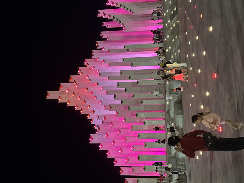 The image depicts an illuminated outdoor plaza at night, specifically at the Ho Chi Minh City Youth Cultural House in District 1, Ho Chi Minh City, Vietnam. The prominent subject is a large, multi-tiered architectural structure composed of numerous vertical rectangular pillars, illuminated internally with vibrant magenta and pink light. The structure's height varies, creating an undulating profile against the dark sky, with each pillar's summit featuring a small, red-glowing element resembling a flame. Below this central structure, multiple individuals are gathered on concrete steps and tiered platforms. In the foreground, a woman wearing a dark red top and dark trousers stands with her back partially to the viewer, holding a mobile phone to her right ear. To her right, a young girl in a light-colored patterned top and shorts is captured in mid-stride. The plaza ground is scattered with numerous small, low-lying lights, predominantly red and yellow. Various other adults and children are observed throughout the scene, standing, walking, or sitting casually.