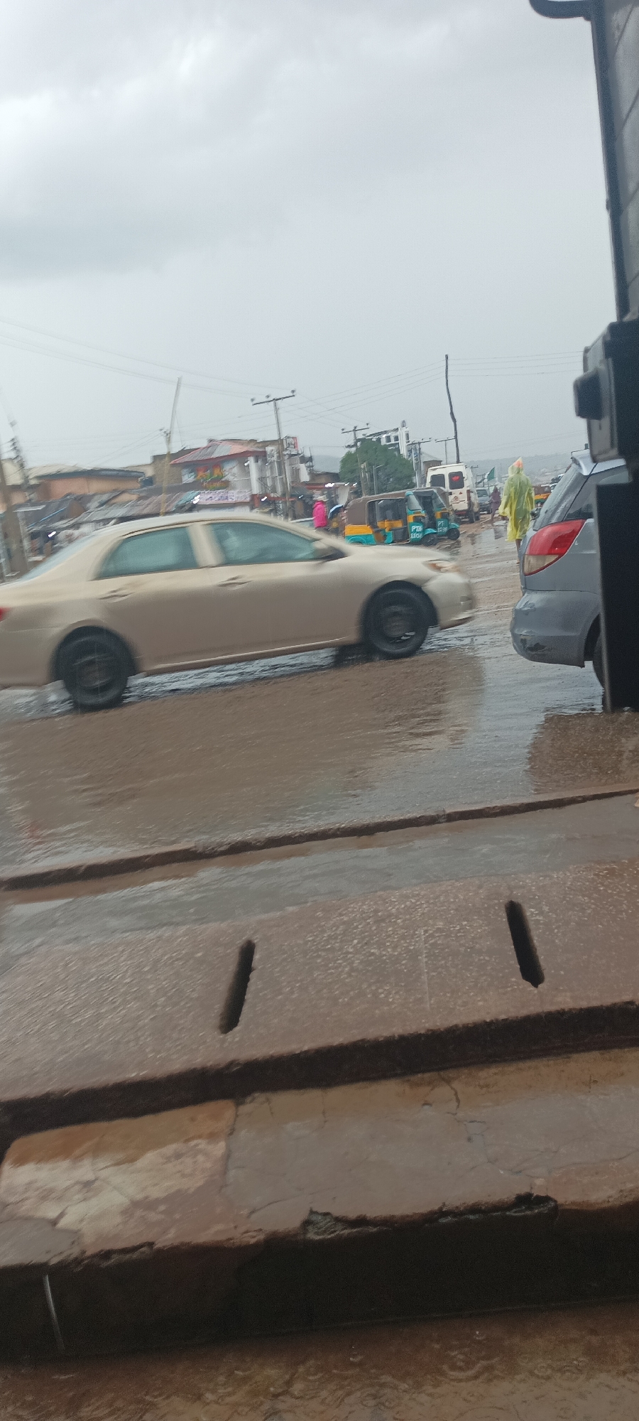 The image captures a rainy or recently rained-on street scene, likely in an urban or semi-urban area, under a heavily overcast, grey sky. The ground is thoroughly wet, with visible puddles and reflections, and what appear to be rain drops disturbing the water in the immediate foreground.
In the mid-ground, a gold-colored sedan, possibly a Toyota Corolla, is moving from left to right, its wheels showing motion blur. To its right, a grey sedan is partially visible and appears stationary. Further back and to the right, a person in a bright yellow rain jacket or poncho is walking. Several Keke NAPEPs (three-wheeled auto-rickshaws, common in Nigeria) are visible in the background, one prominently featuring blue and yellow colors. A white van is also present further down the road.
The background features various buildings, some appearing like shops or residential structures, with power poles and overhead electrical wires crisscrossing the sky. The architecture and vehicle types suggest a setting in a developing country, possibly West Africa.
In the immediate foreground, there's a concrete or paved surface, possibly a sidewalk or a drainage channel, with rectangular cutouts functioning as storm drains. This surface is also wet and shows signs of wear and cracking. On one of the Keke NAPEPs, partial text "PTM" and "LOG" can be seen, likely part of a company or service name such as "PTM LOGISTICS". The overall atmosphere is muted due to the grey weather conditions.