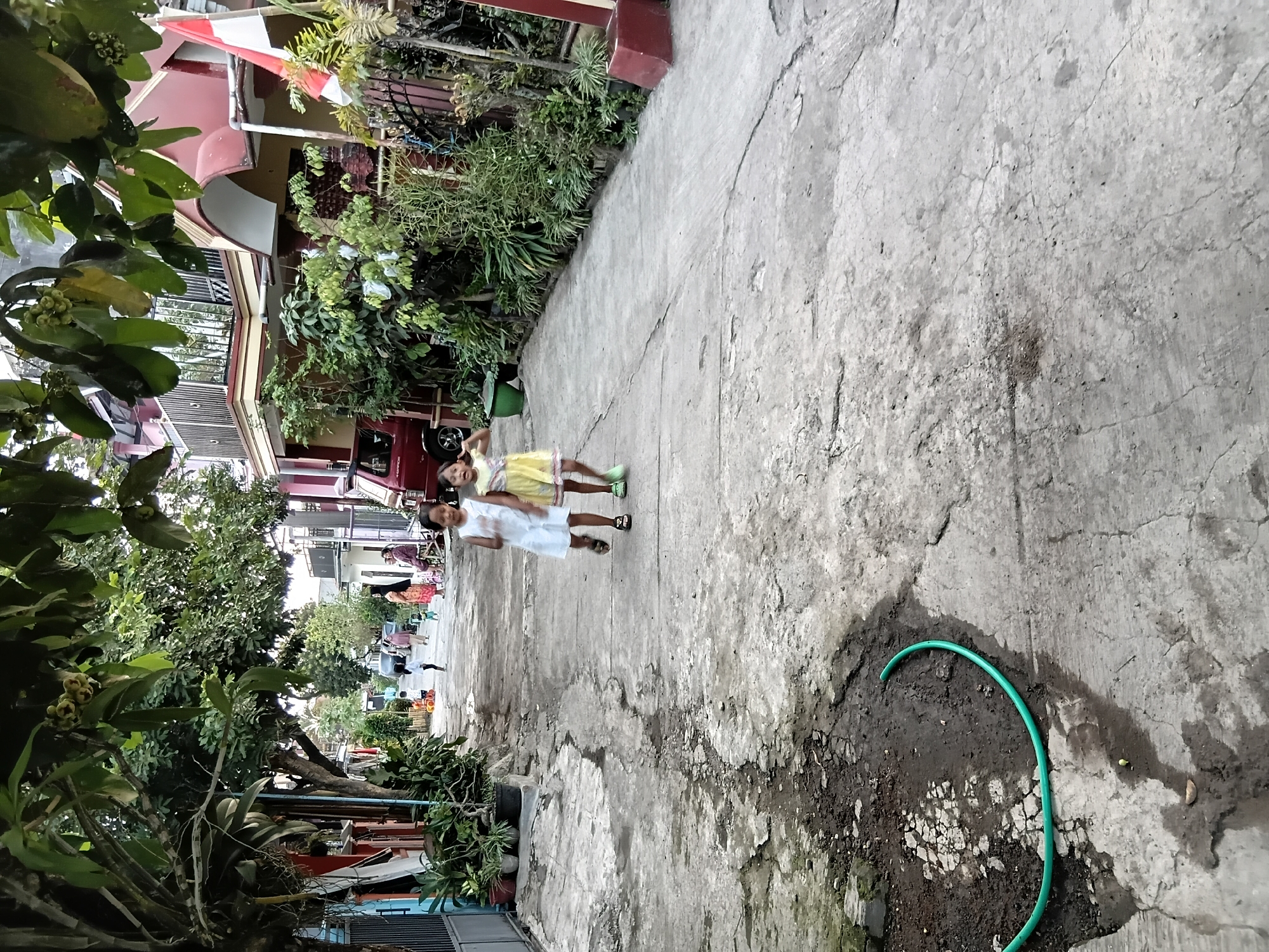 The image depicts a narrow, paved alleyway in Tanggul Wetan, Indonesia, lined with residential buildings and dense vegetation. Two young girls, one in a white dress and the other in a yellow dress, are running towards the camera in the middle of the alley. The girl in white wears dark sandals, and the girl in yellow wears green sandals. In the background, other people are visible, including adults and children, and a red vehicle is parked on the side. A green garden hose lies coiled on the cracked concrete pavement in the foreground, near a patch of dark, muddy ground. The buildings display red and white facades, with balconies and foliage. An Indonesian flag is prominently displayed on a pole on the right side of the frame. The overall impression is that of a residential neighborhood with active outdoor life.