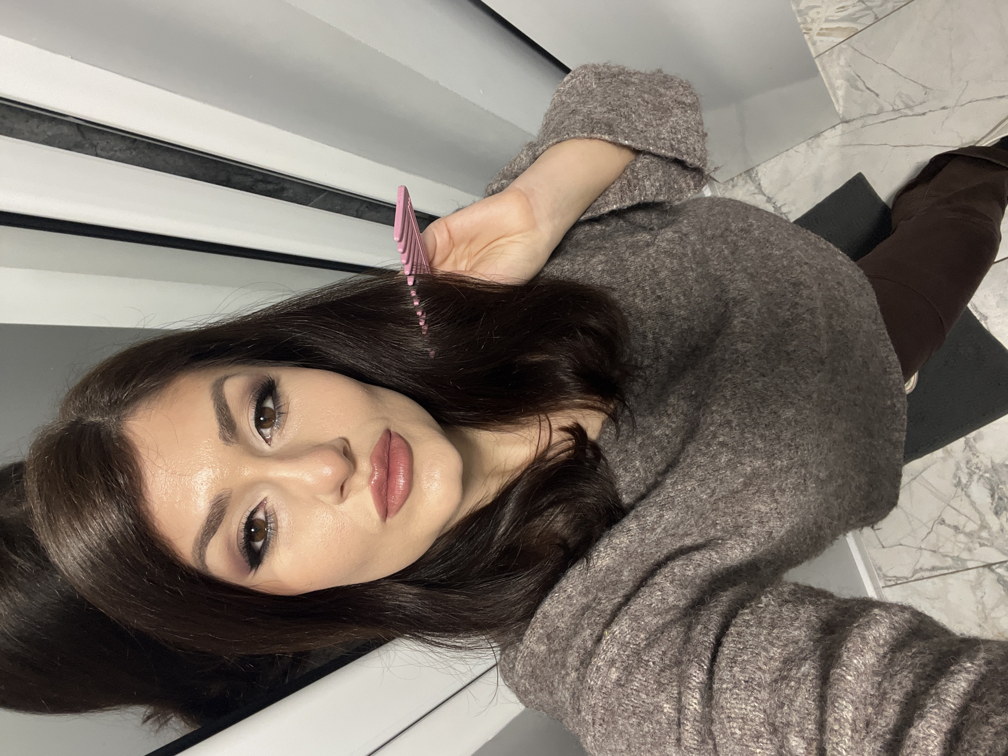 A woman with dark brown hair, light skin, and wearing a grey sweater is holding a pink comb near her hair. She appears to be indoors, as indicated by the wall behind her which is partially white with dark grey stripes. The floor beneath her is marble-patterned. A dark object, possibly a chair or bag, is visible to the right of the woman. There is no indication of the location being the city of Yambol, Bulgaria.