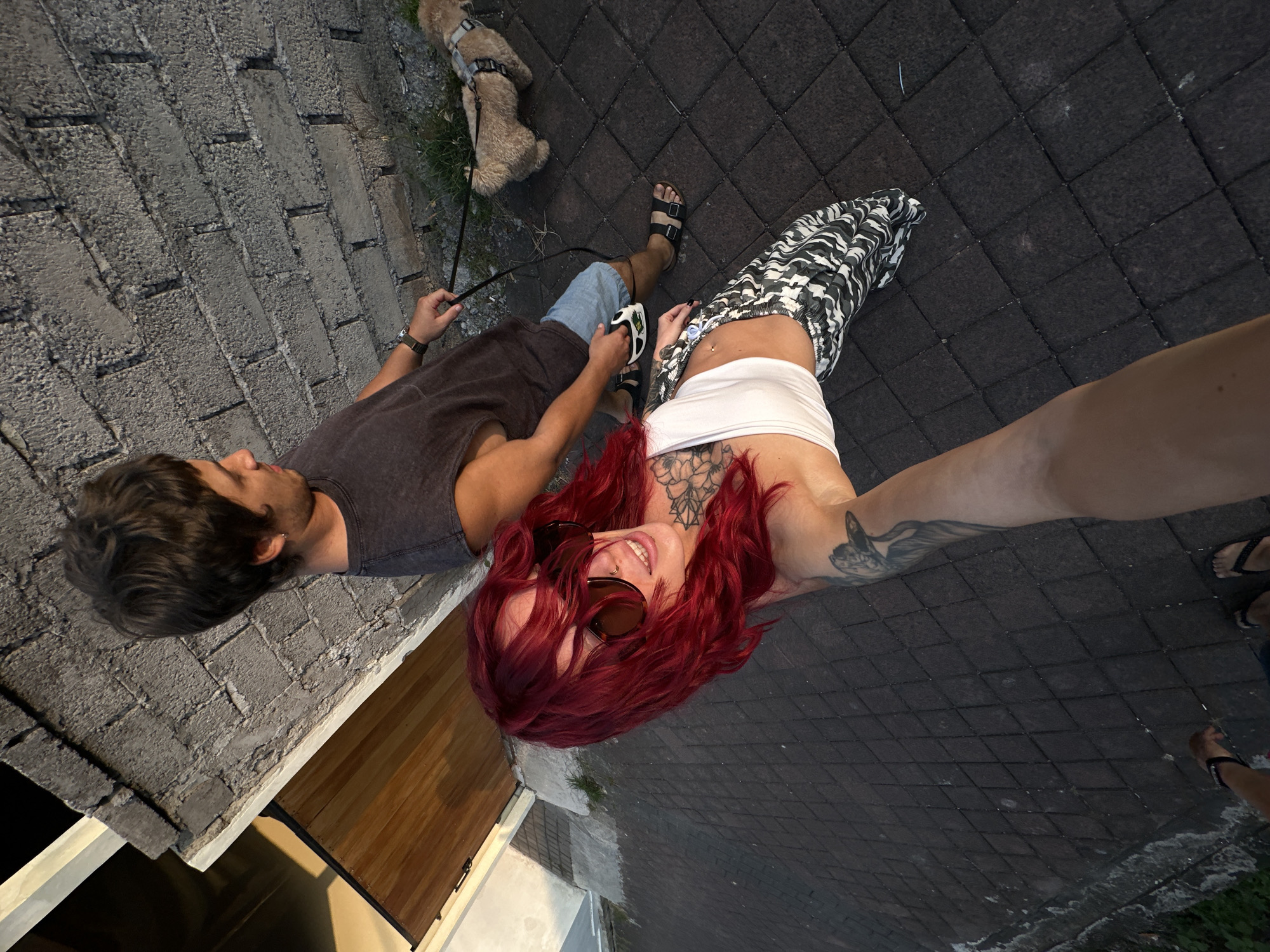 In North Kuta, Indonesia, a man and a woman are walking a dog on a brick-paved path. The man, wearing a dark t-shirt and blue shorts, holds the dog's leash. The woman, with bright red hair and sunglasses, wears a white crop top and patterned pants. She has tattoos on her arm and chest. A small, light-colored dog on a harness walks beside the man. The background features a textured brick wall and a wooden door. The scene is illuminated by late afternoon sunlight.